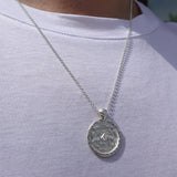 A silver chain necklace with a hammered circular pendant featuring a small central stone, worn on a person's neck over a white t-shirt.