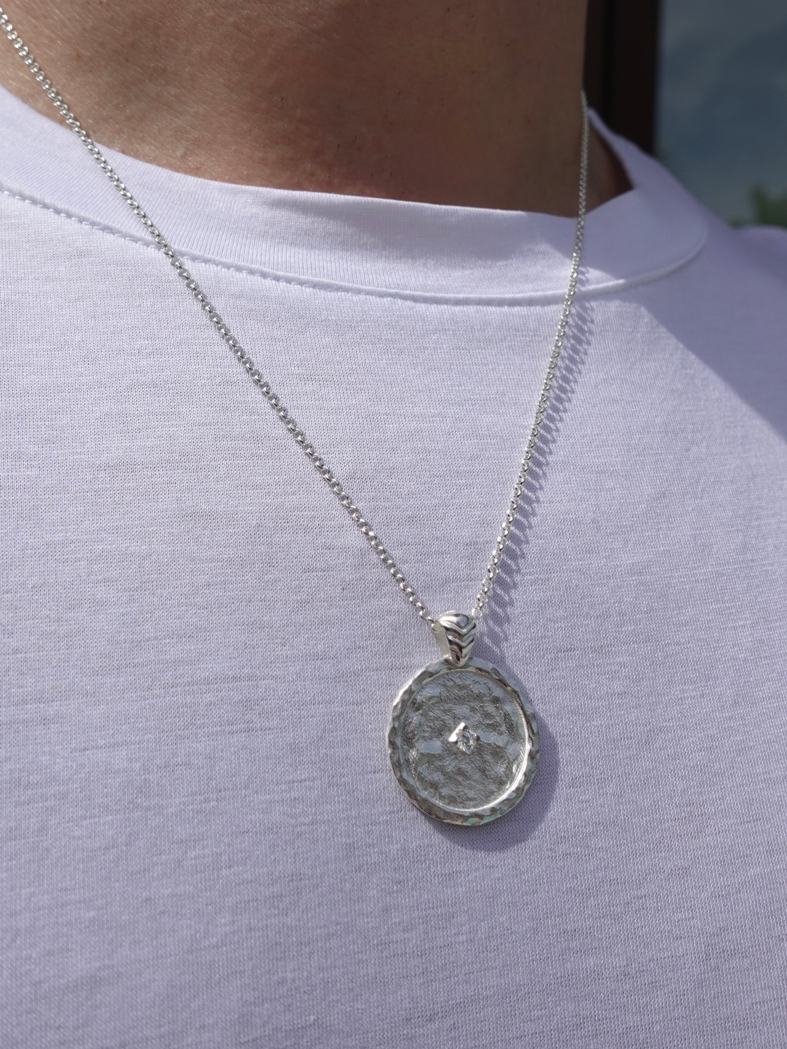 A silver chain necklace with a hammered circular pendant featuring a small central stone, worn on a person's neck over a white t-shirt.