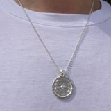 A silver pendant necklace with a textured circular medallion pendant, shown worn on a person's neck over a white t-shirt.