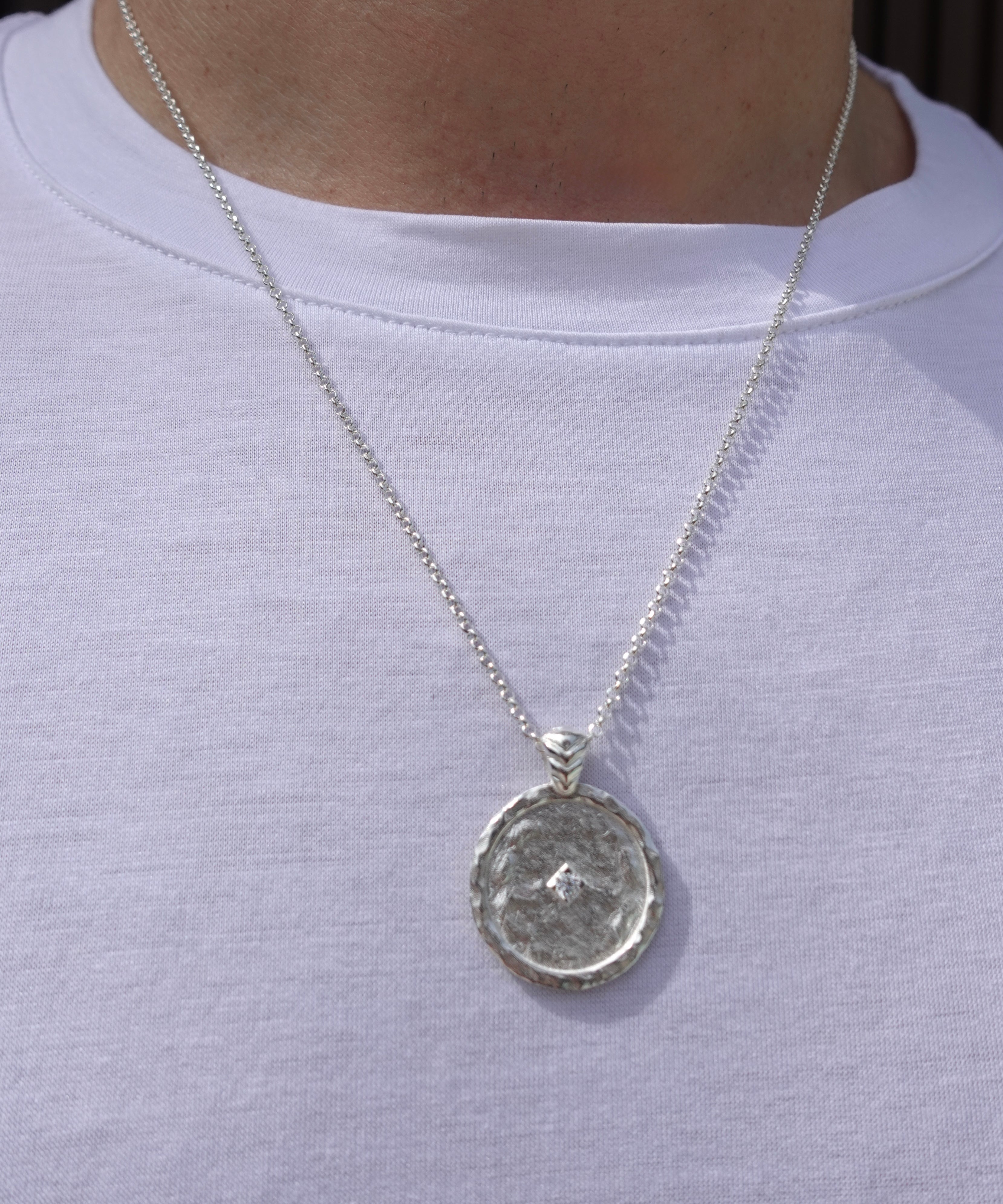 A silver pendant necklace with a textured circular medallion pendant, shown worn on a person's neck over a white t-shirt.