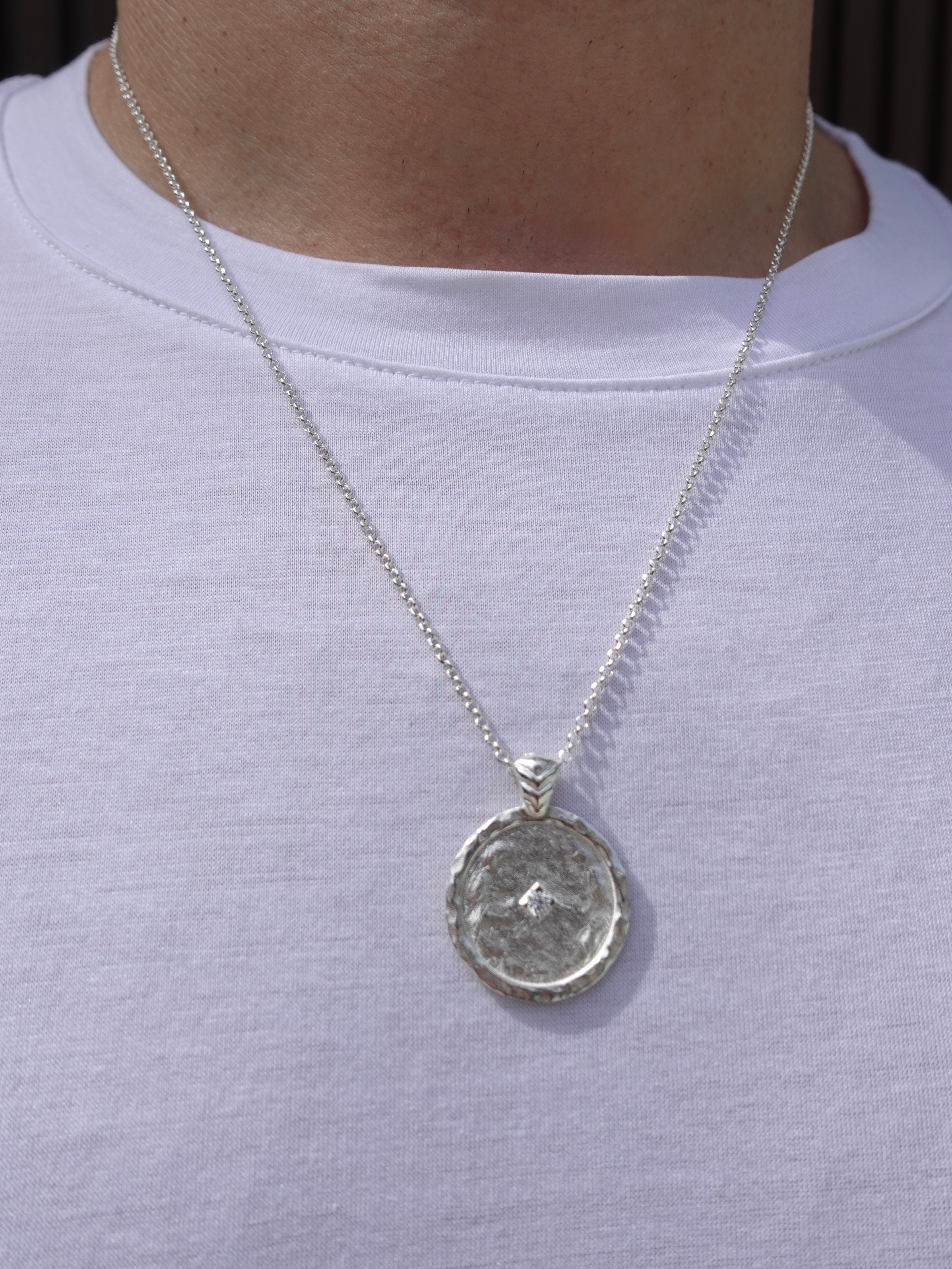 A silver pendant necklace with a textured circular medallion pendant, shown worn on a person's neck over a white t-shirt.