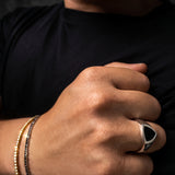 A close-up of a man's hand wearing a silver signet ring with a black triangular face, alongside two beaded bracelets, against a dark background.