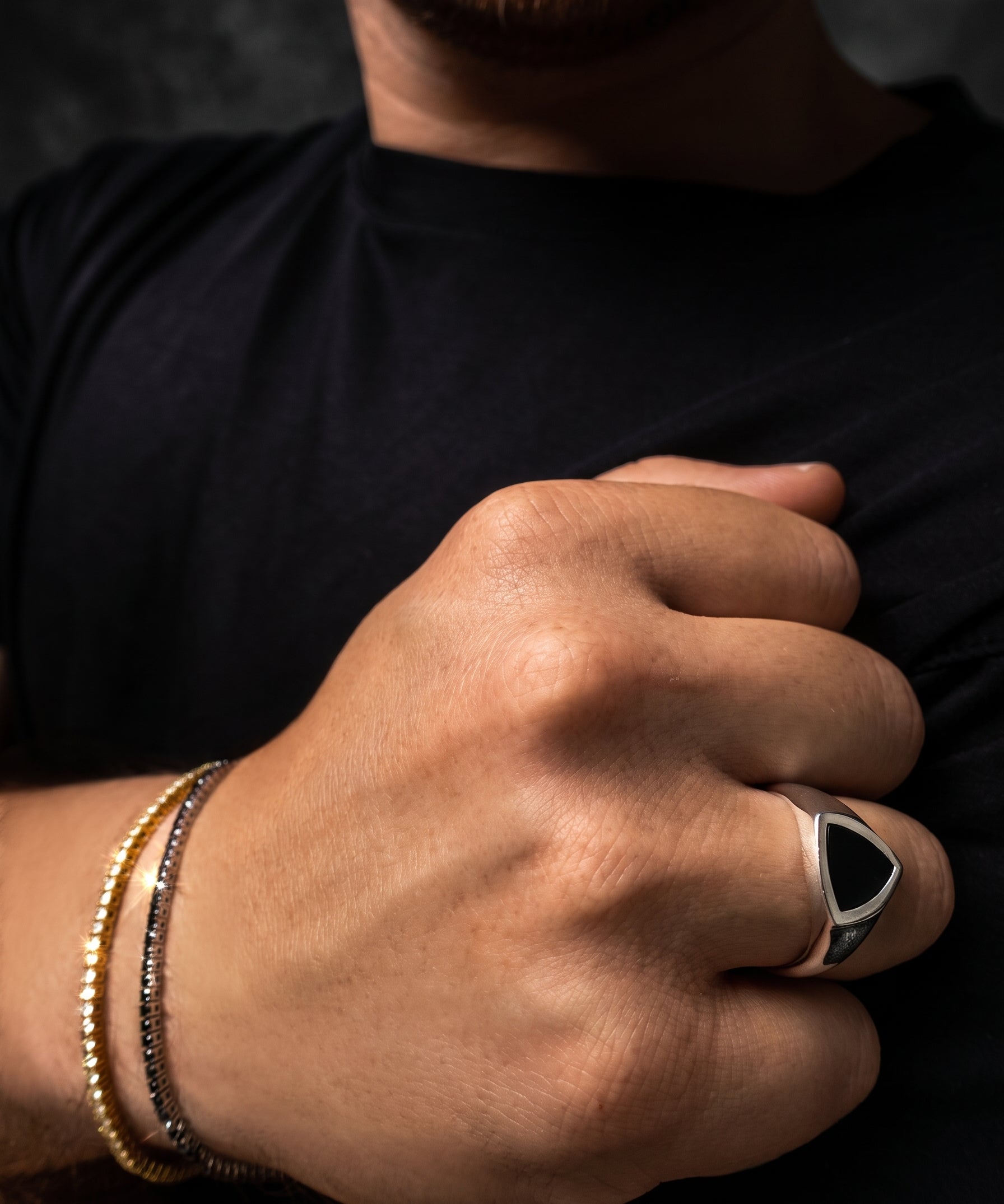 A close-up of a man's hand wearing a silver signet ring with a black triangular face, alongside two beaded bracelets, against a dark background.