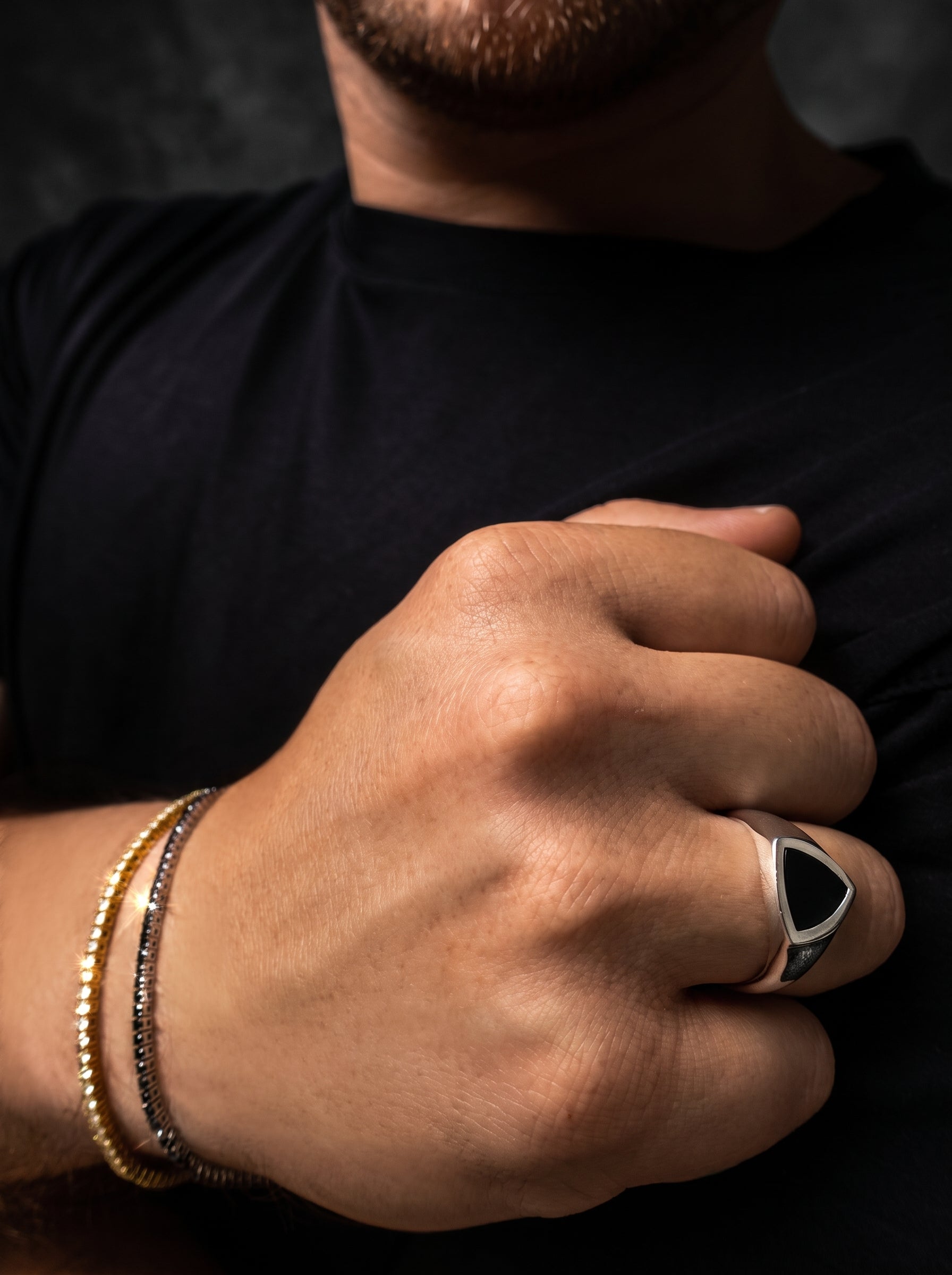 A close-up of a man's hand wearing a silver signet ring with a black triangular face, alongside two beaded bracelets, against a dark background.