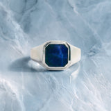 A silver signet ring with a faceted blue stone, photographed on a light blue marble surface.