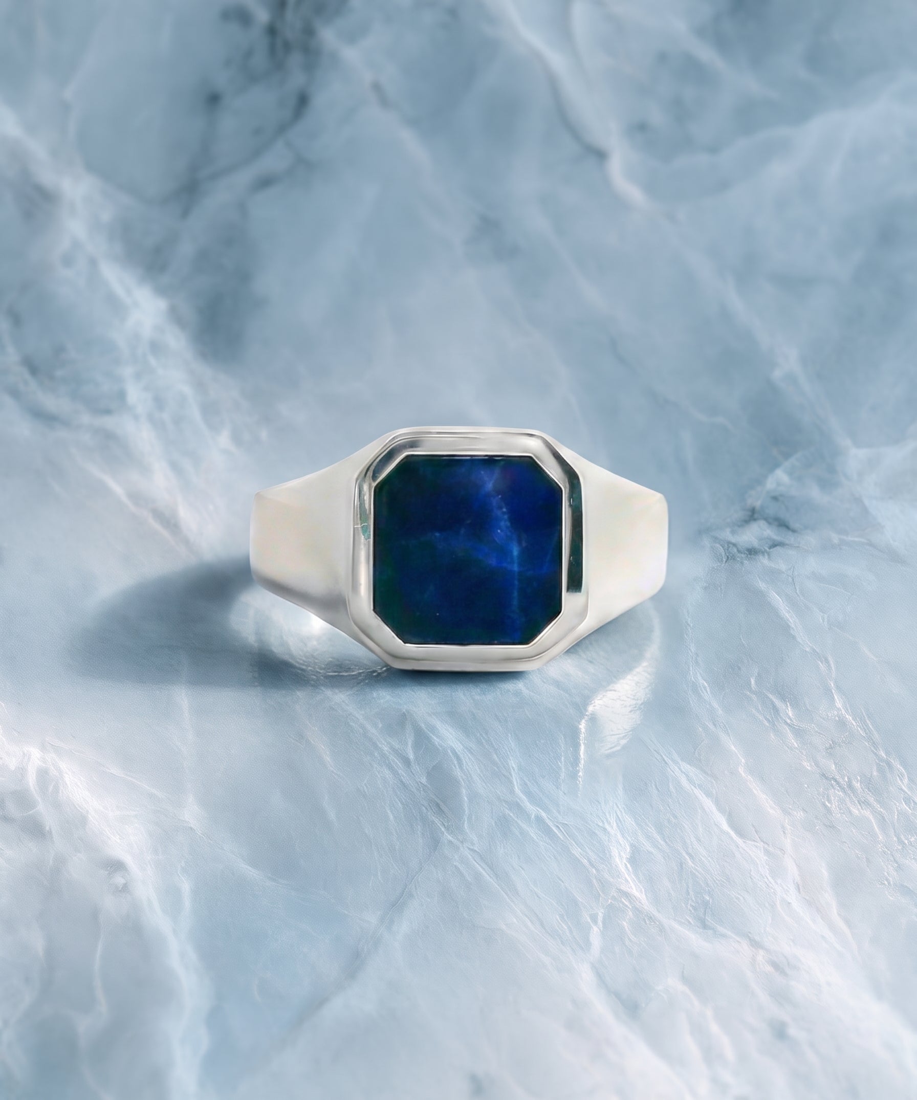 A silver signet ring with a faceted blue stone, photographed on a light blue marble surface.