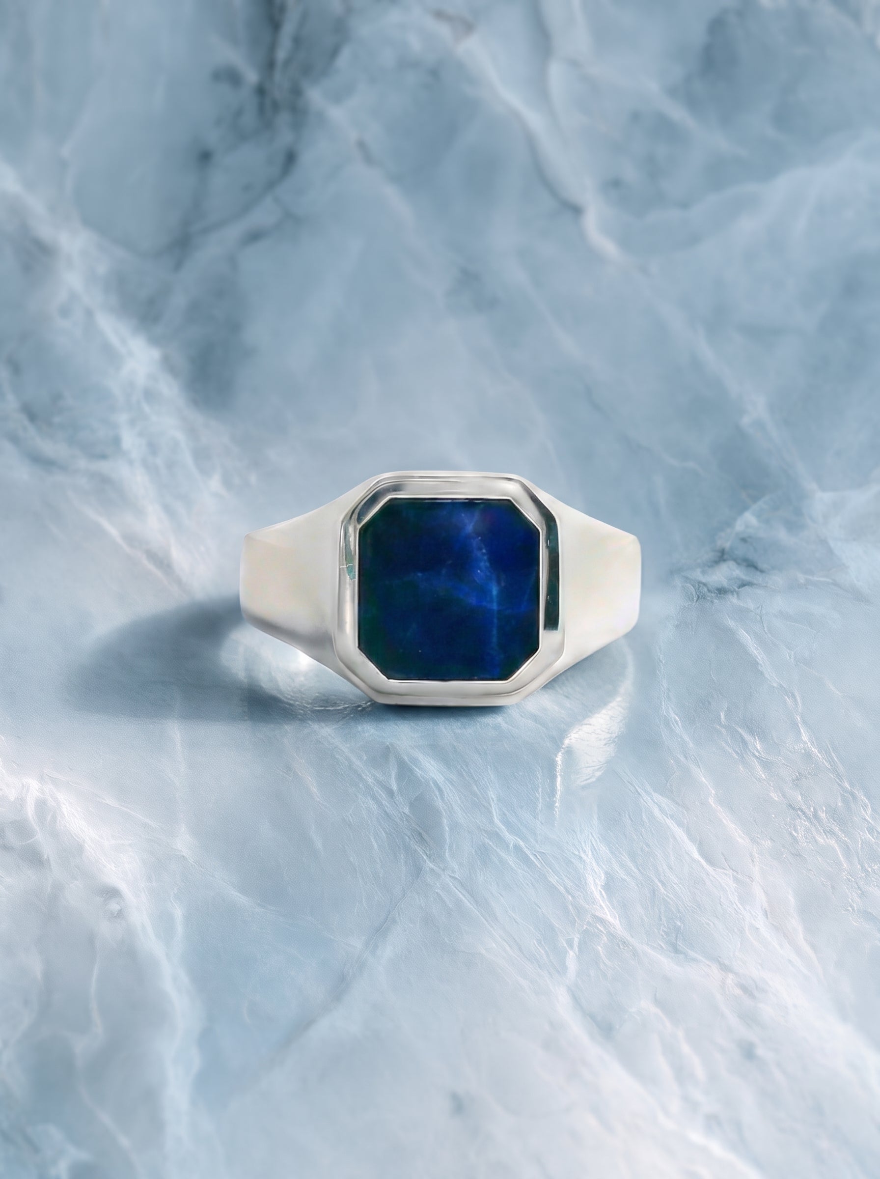 A silver signet ring with a faceted blue stone, photographed on a light blue marble surface.