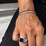 A close-up of a person's hand wearing a silver-toned signet ring with a large blue stone and two delicate silver bracelets, one set with dark blue stones and the other with clear stones.