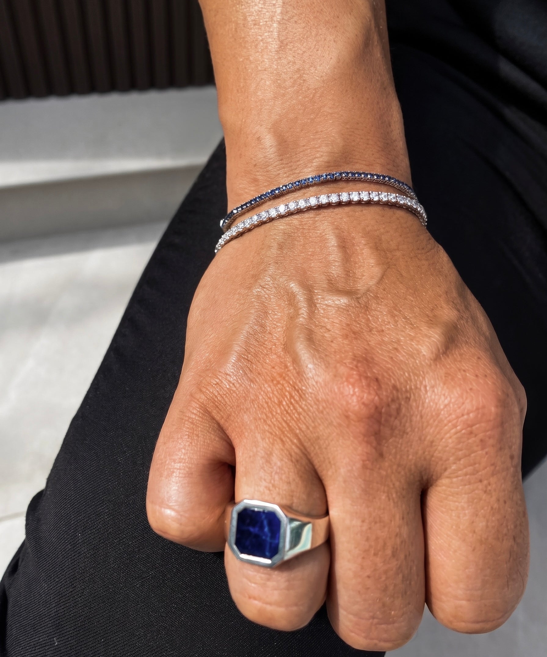 A close-up of a person's hand wearing a silver-toned signet ring with a large blue stone and two delicate silver bracelets, one set with dark blue stones and the other with clear stones.