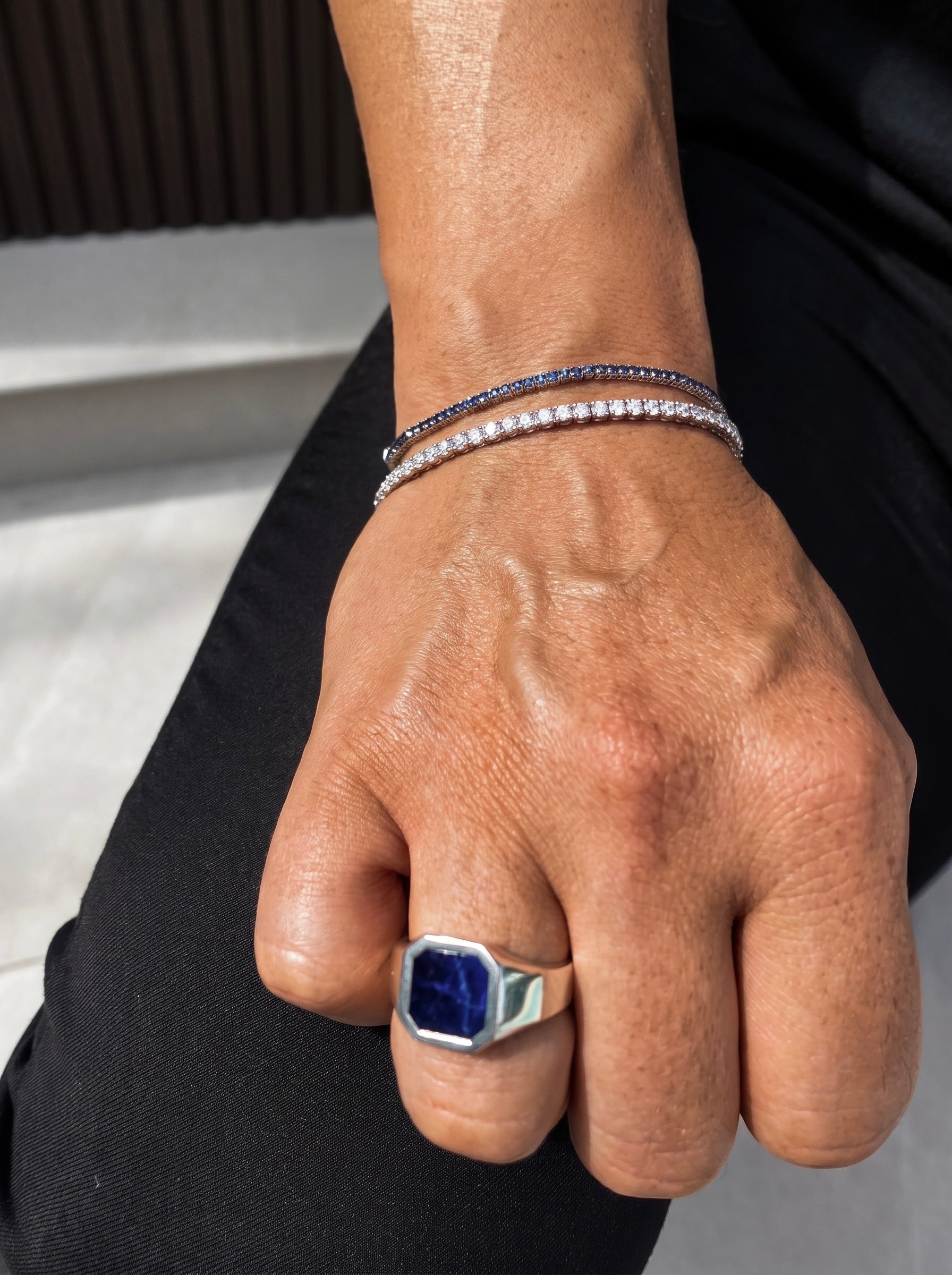 A close-up of a person's hand wearing a silver-toned signet ring with a large blue stone and two delicate silver bracelets, one set with dark blue stones and the other with clear stones.
