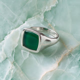 A silver signet ring with a square-cut dark green stone, displayed on a light green marbled surface.