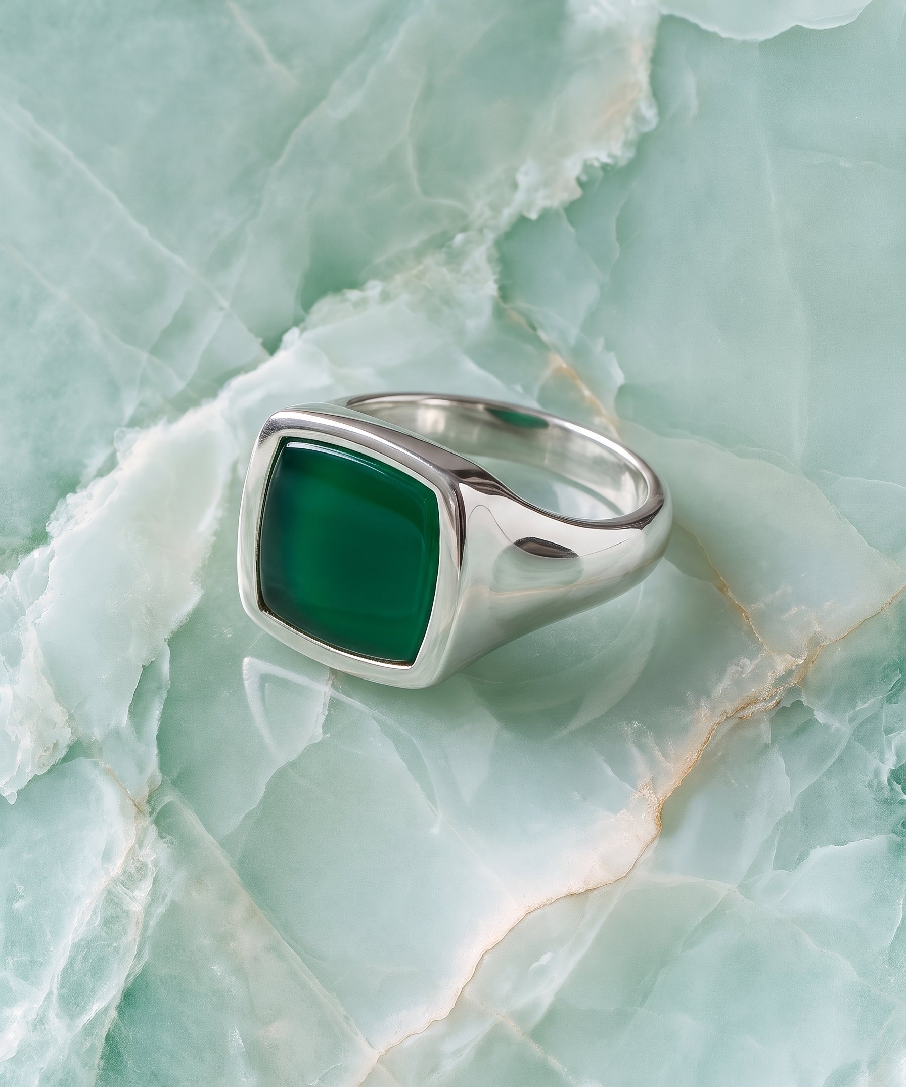 A silver signet ring with a square-cut dark green stone, displayed on a light green marbled surface.