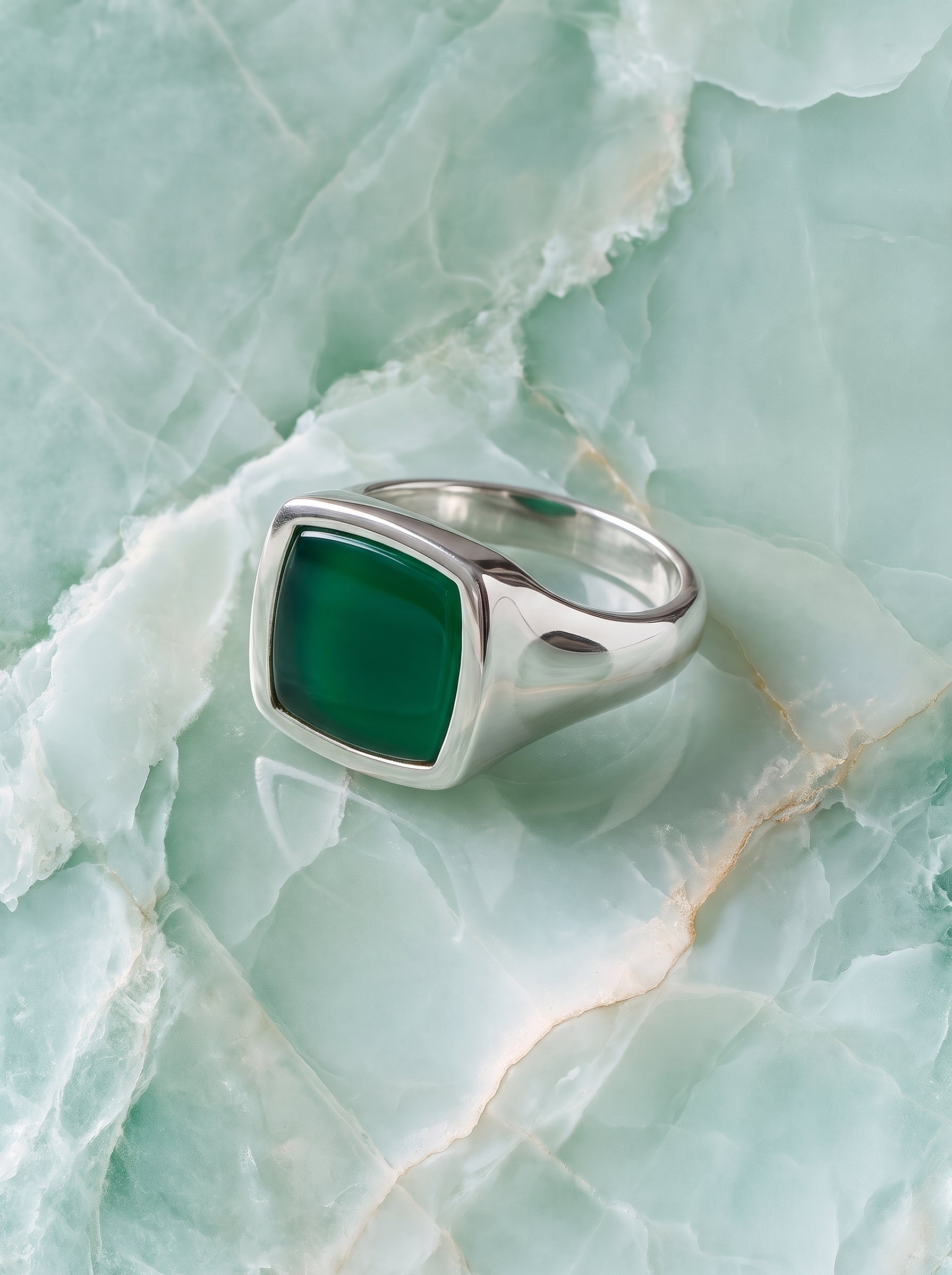 A silver signet ring with a square-cut dark green stone, displayed on a light green marbled surface.