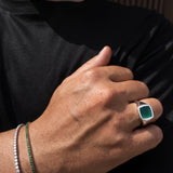 A close-up lifestyle shot of a person's hand wearing a silver signet ring with a square green cabochon stone, alongside a green beaded bracelet and a silver beaded bracelet on the wrist, set against a black t-shirt.