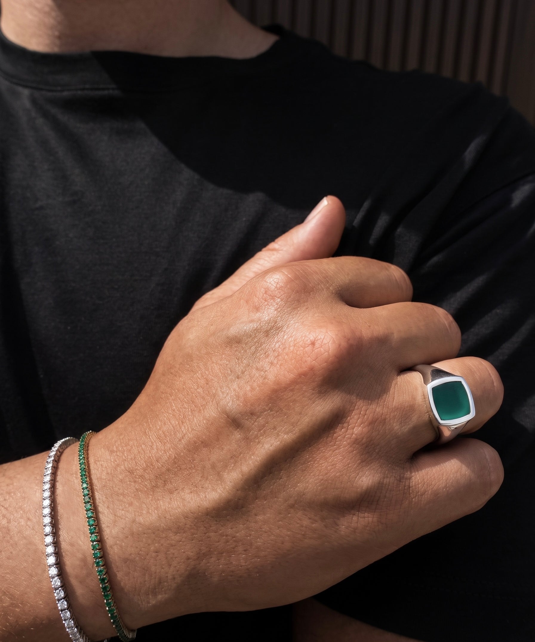 A close-up lifestyle shot of a person's hand wearing a silver signet ring with a square green cabochon stone, alongside a green beaded bracelet and a silver beaded bracelet on the wrist, set against a black t-shirt.