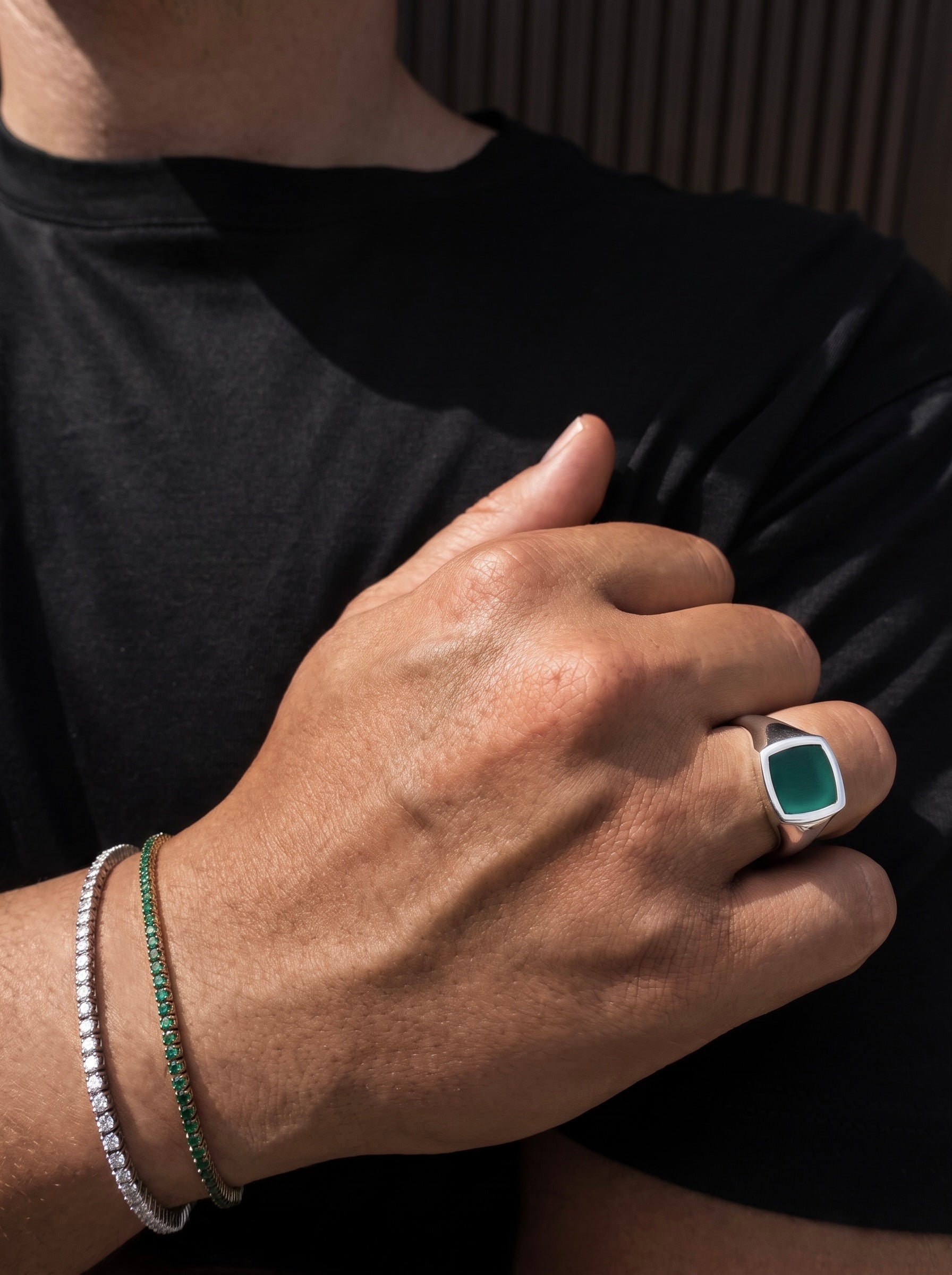 A close-up lifestyle shot of a person's hand wearing a silver signet ring with a square green cabochon stone, alongside a green beaded bracelet and a silver beaded bracelet on the wrist, set against a black t-shirt.