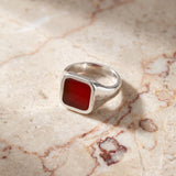 A silver signet ring with a square red stone, resting on a beige marble surface with soft shadows.