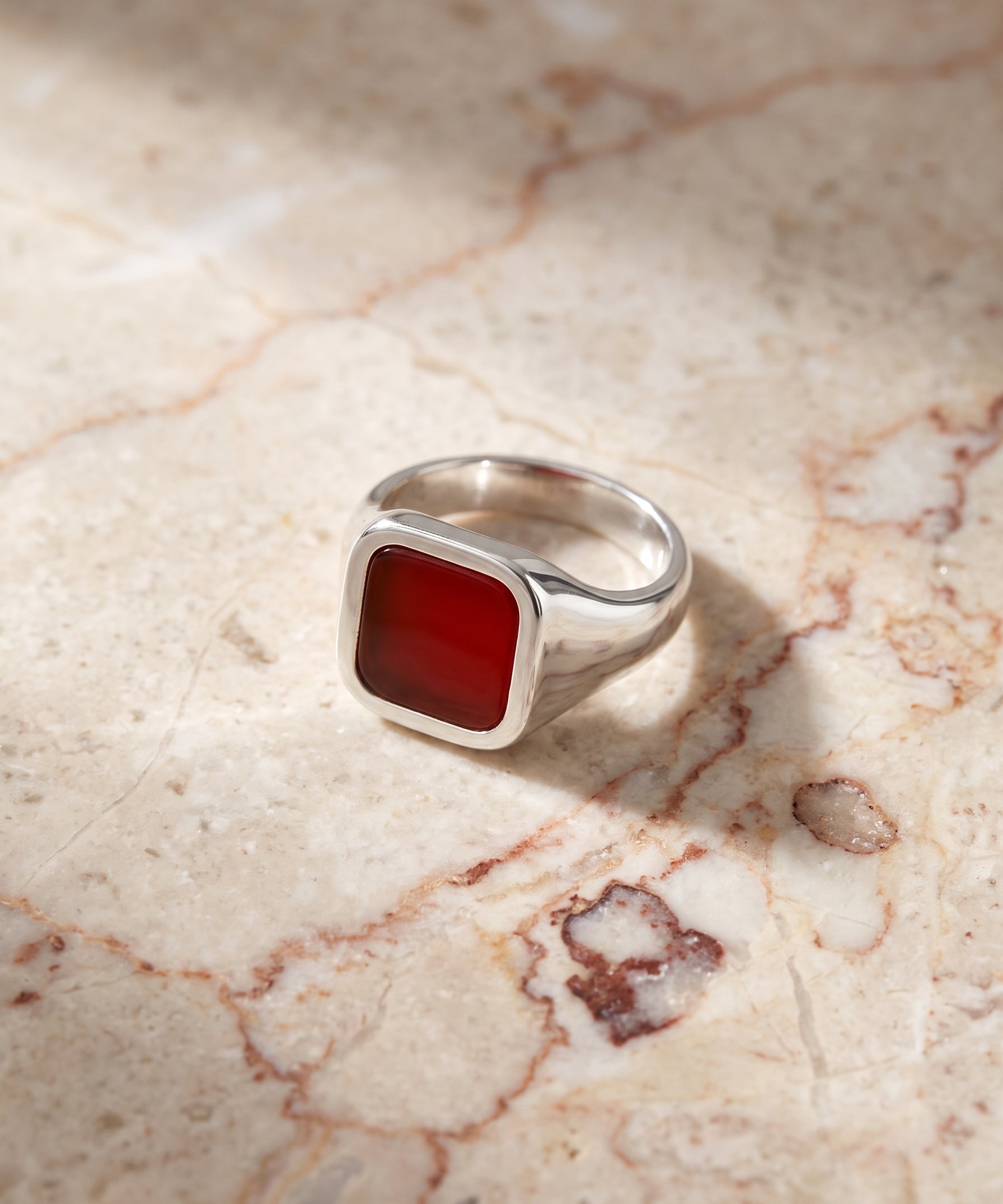 A silver signet ring with a square red stone, resting on a beige marble surface with soft shadows.