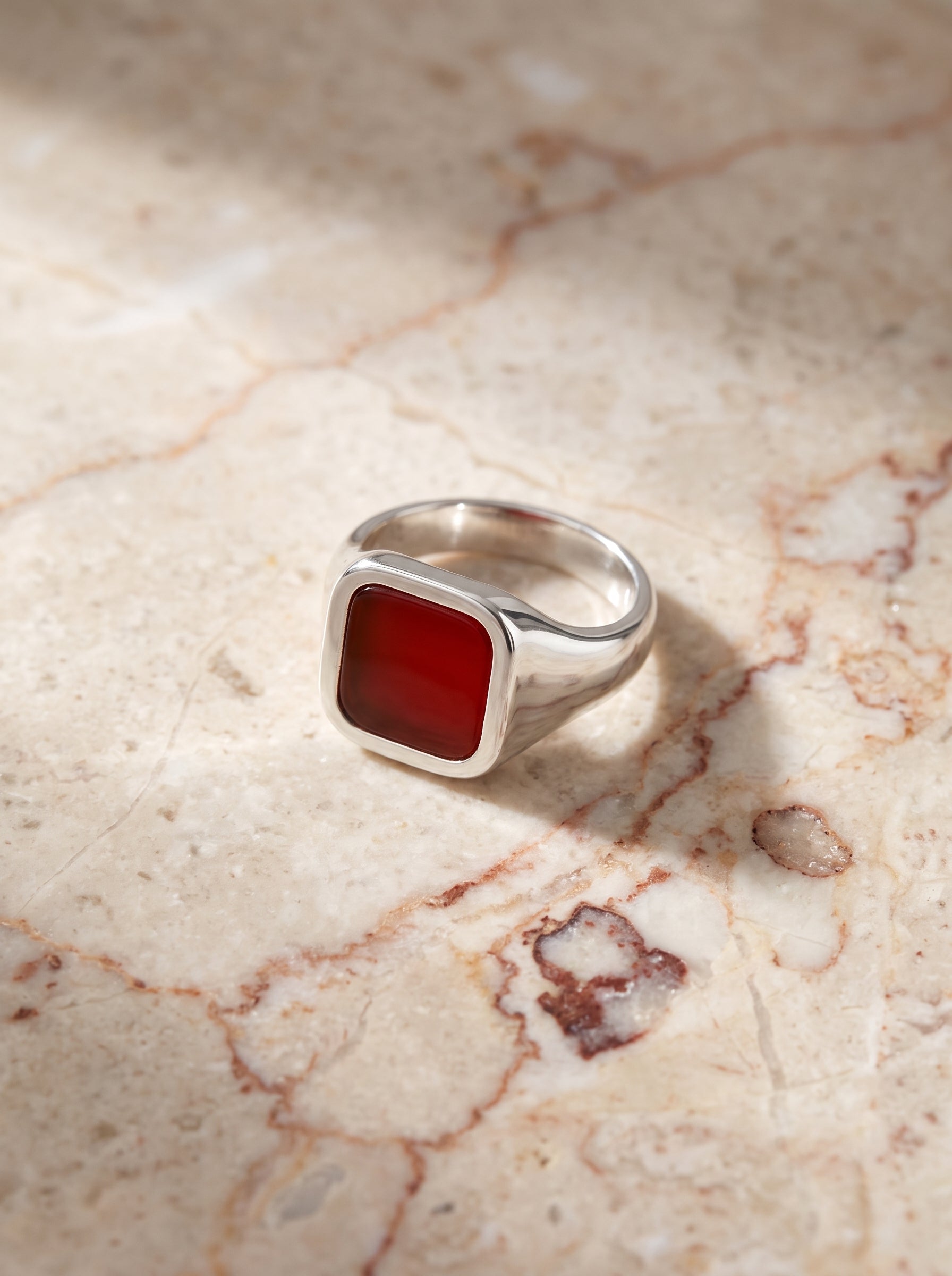A silver signet ring with a square red stone, resting on a beige marble surface with soft shadows.