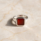 A silver signet ring with a square, faceted red gemstone, resting on a light beige marble surface.