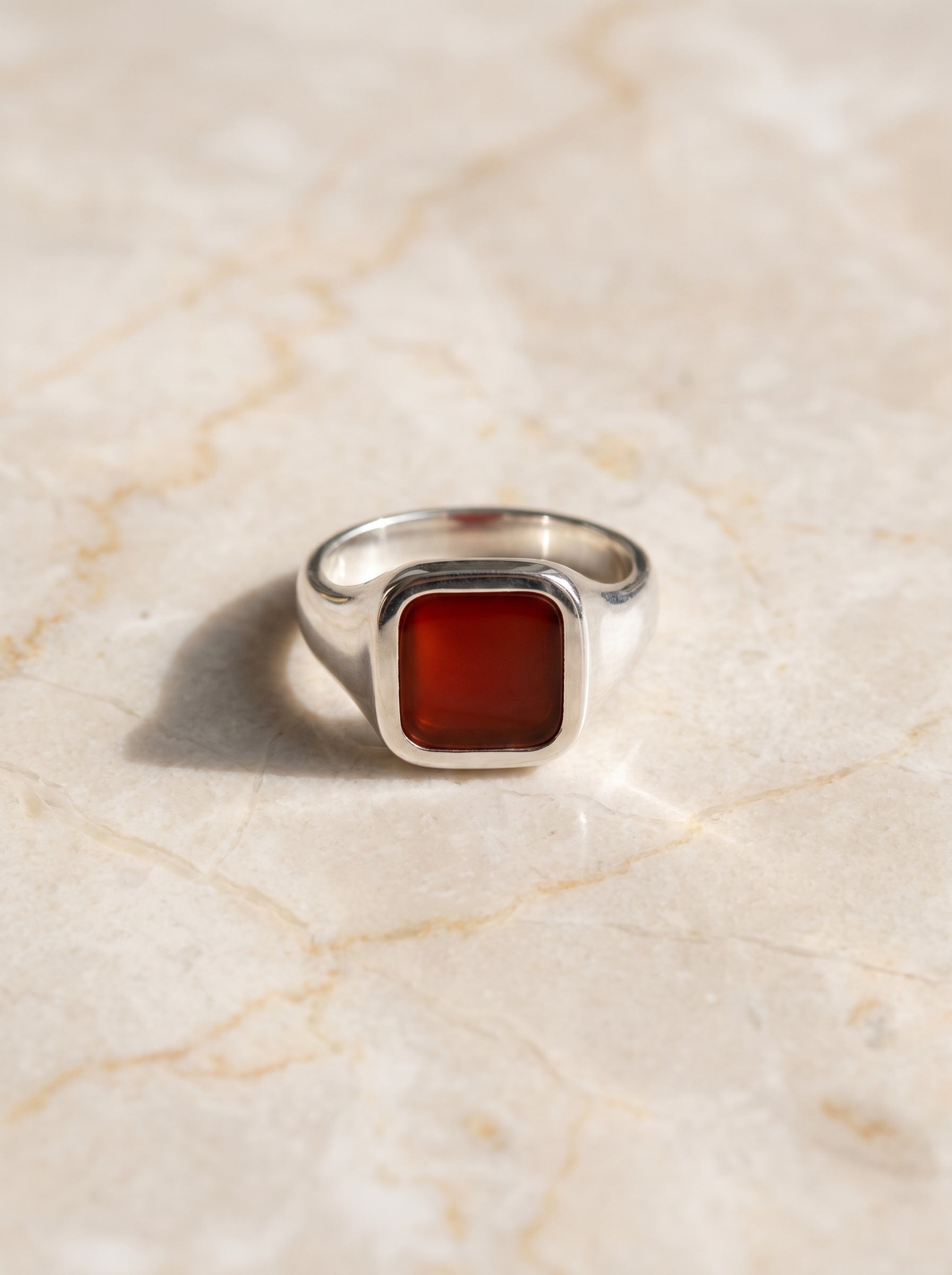 A silver signet ring with a square, faceted red gemstone, resting on a light beige marble surface.