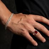 A close-up of a person's hand resting on their hip, showcasing a silver signet ring with a square red gemstone and a silver triple-band bracelet.