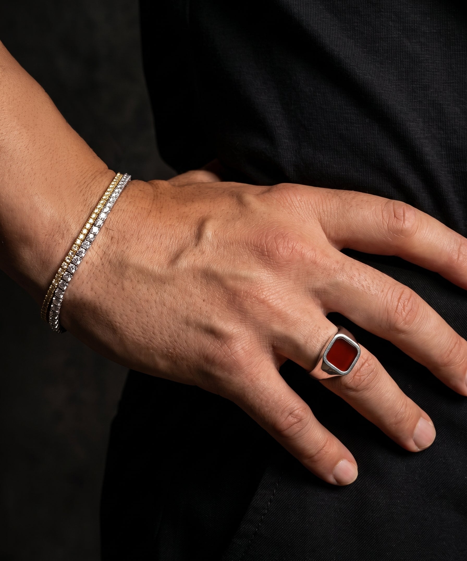 A close-up of a person's hand resting on their hip, showcasing a silver signet ring with a square red gemstone and a silver triple-band bracelet.