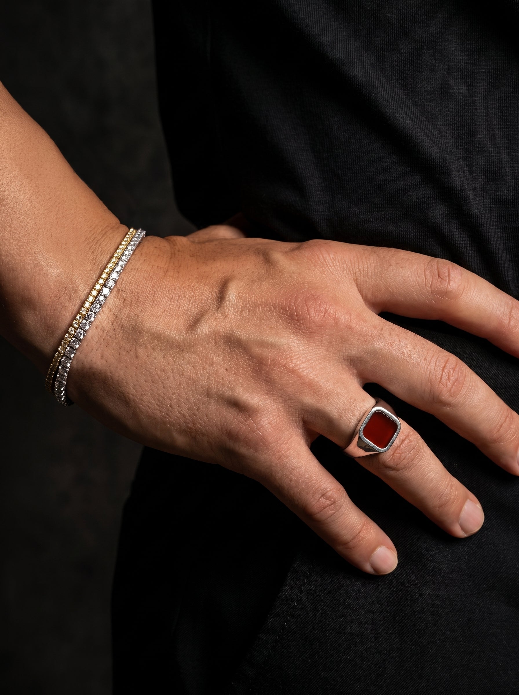 A close-up of a person's hand resting on their hip, showcasing a silver signet ring with a square red gemstone and a silver triple-band bracelet.
