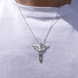 A silver pendant necklace featuring a stylized dagger with wings, worn on a person's neck over a white t-shirt.
