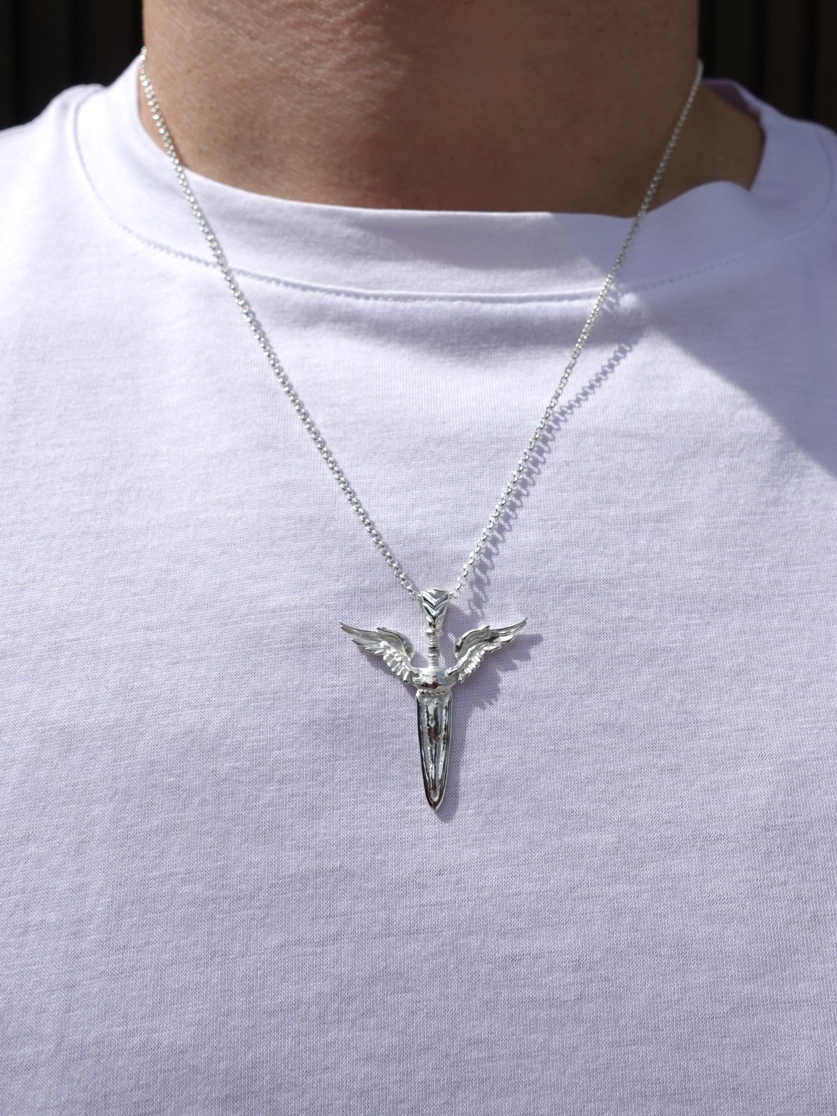 A silver pendant necklace featuring a stylized dagger with wings, worn on a person's neck over a white t-shirt.