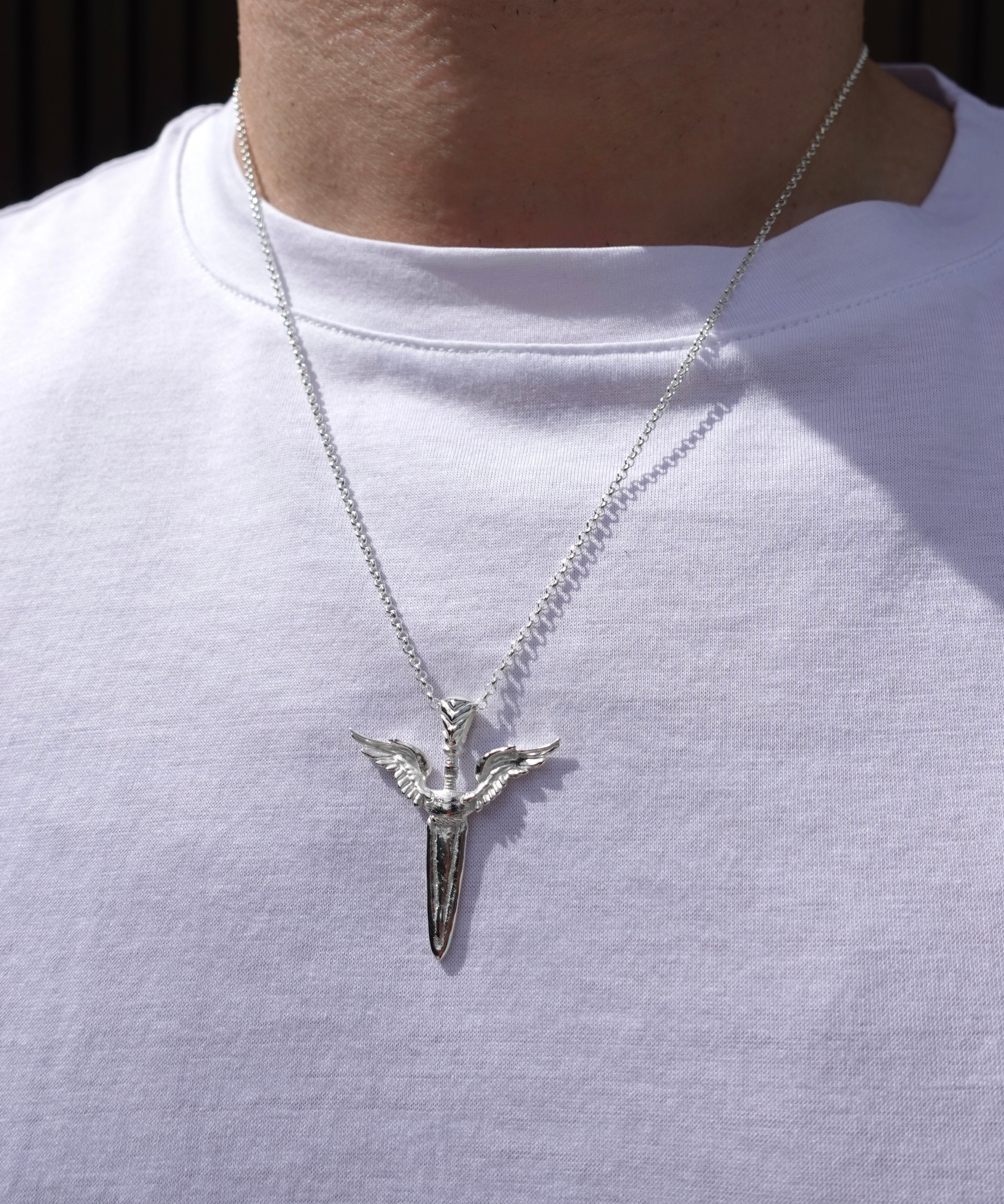 A silver pendant necklace featuring a stylized winged sword emblem on a chain, worn by a person wearing a white t-shirt.
