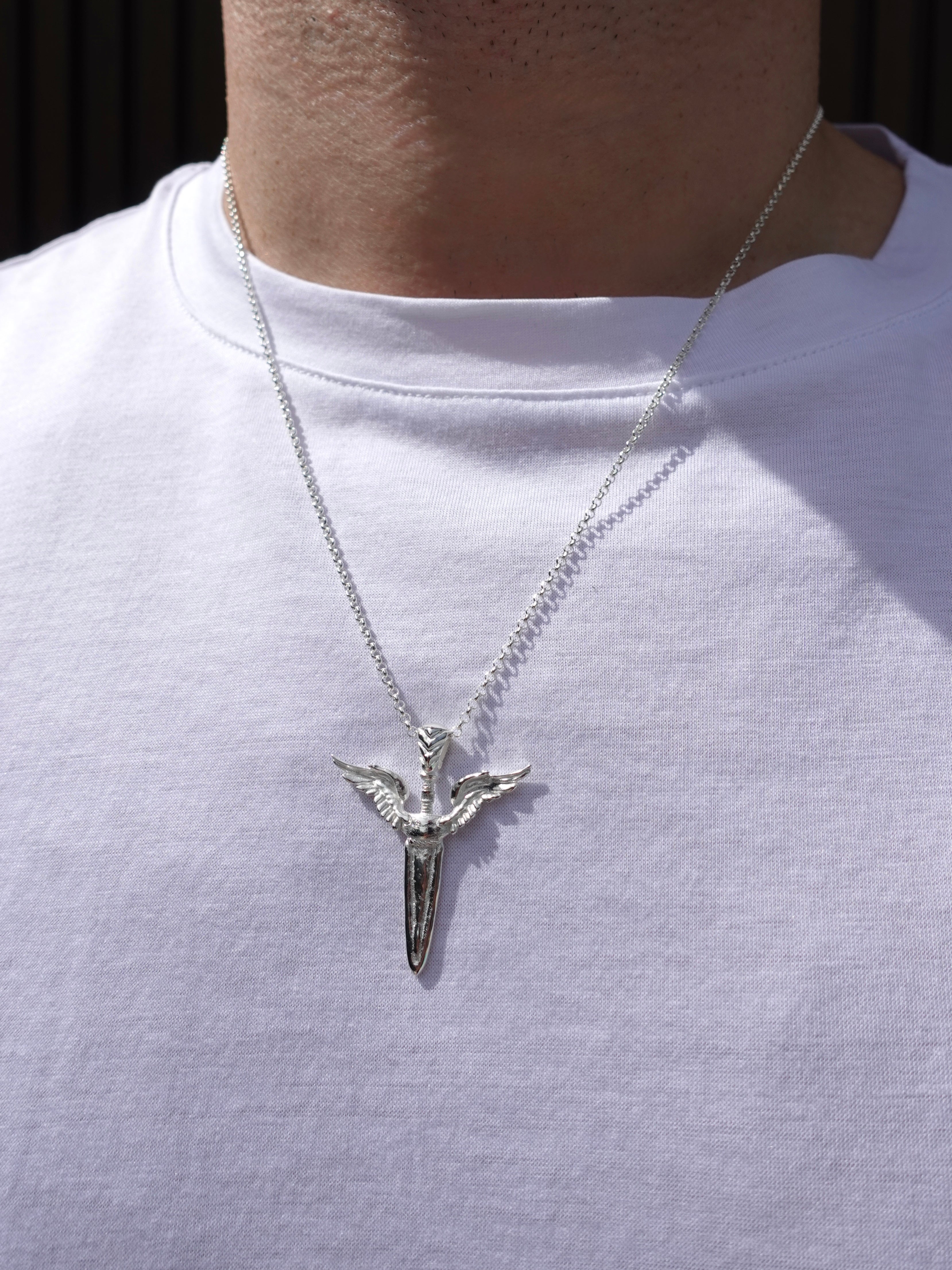 A silver pendant necklace featuring a stylized winged sword emblem on a chain, worn by a person wearing a white t-shirt.