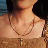 Close-up of a person wearing a gold necklace with a winged-shaped sword pendant against a blurred natural background