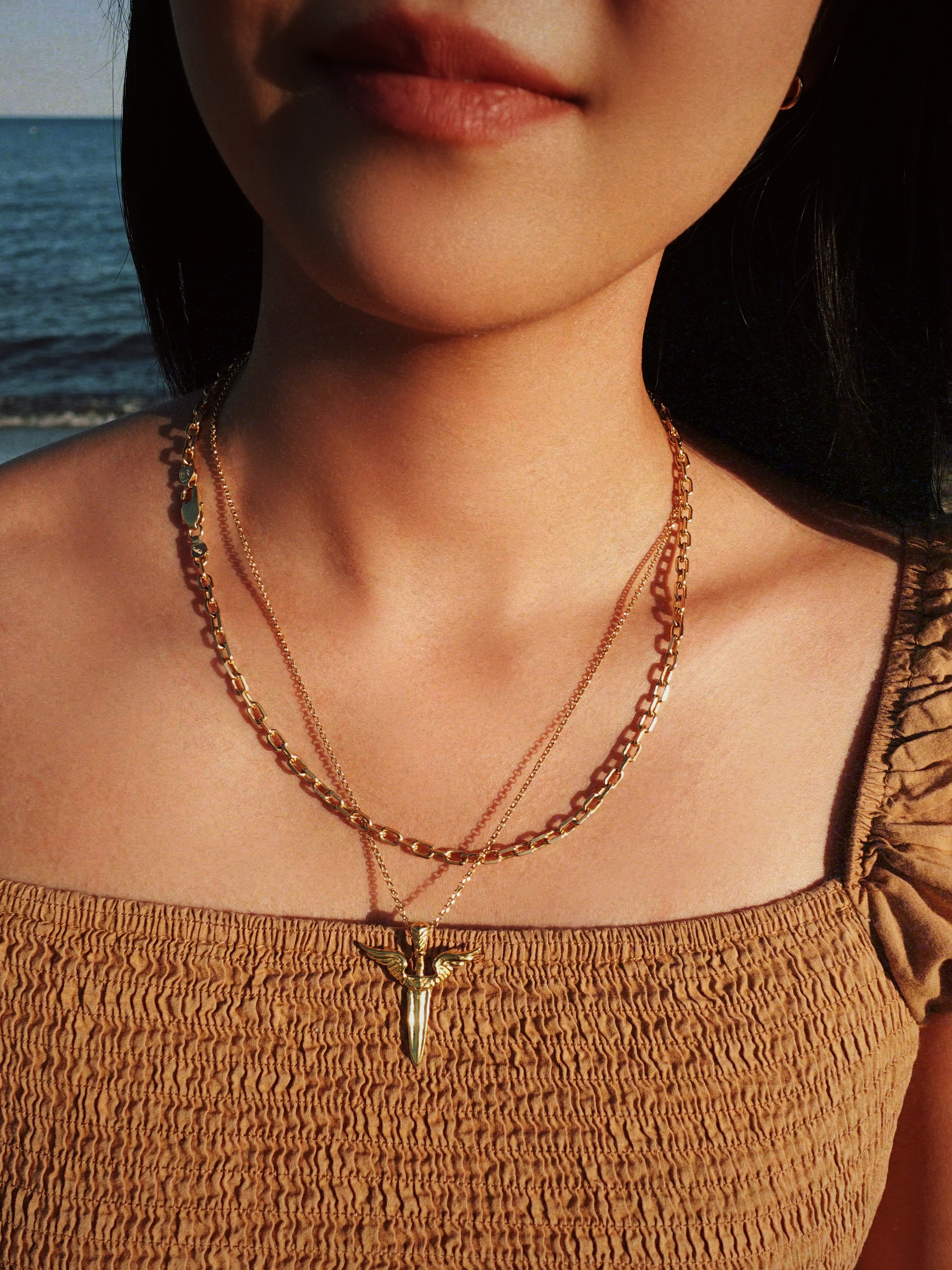 Close-up of a person wearing a gold necklace with a winged-shaped sword pendant against a blurred natural background
