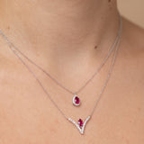 A close-up of a model's neck and collarbone wearing two layered silver necklaces. The shorter necklace features a teardrop-shaped ruby pendant, while the longer necklace has a V-shaped pendant with a small ruby and clear stones.