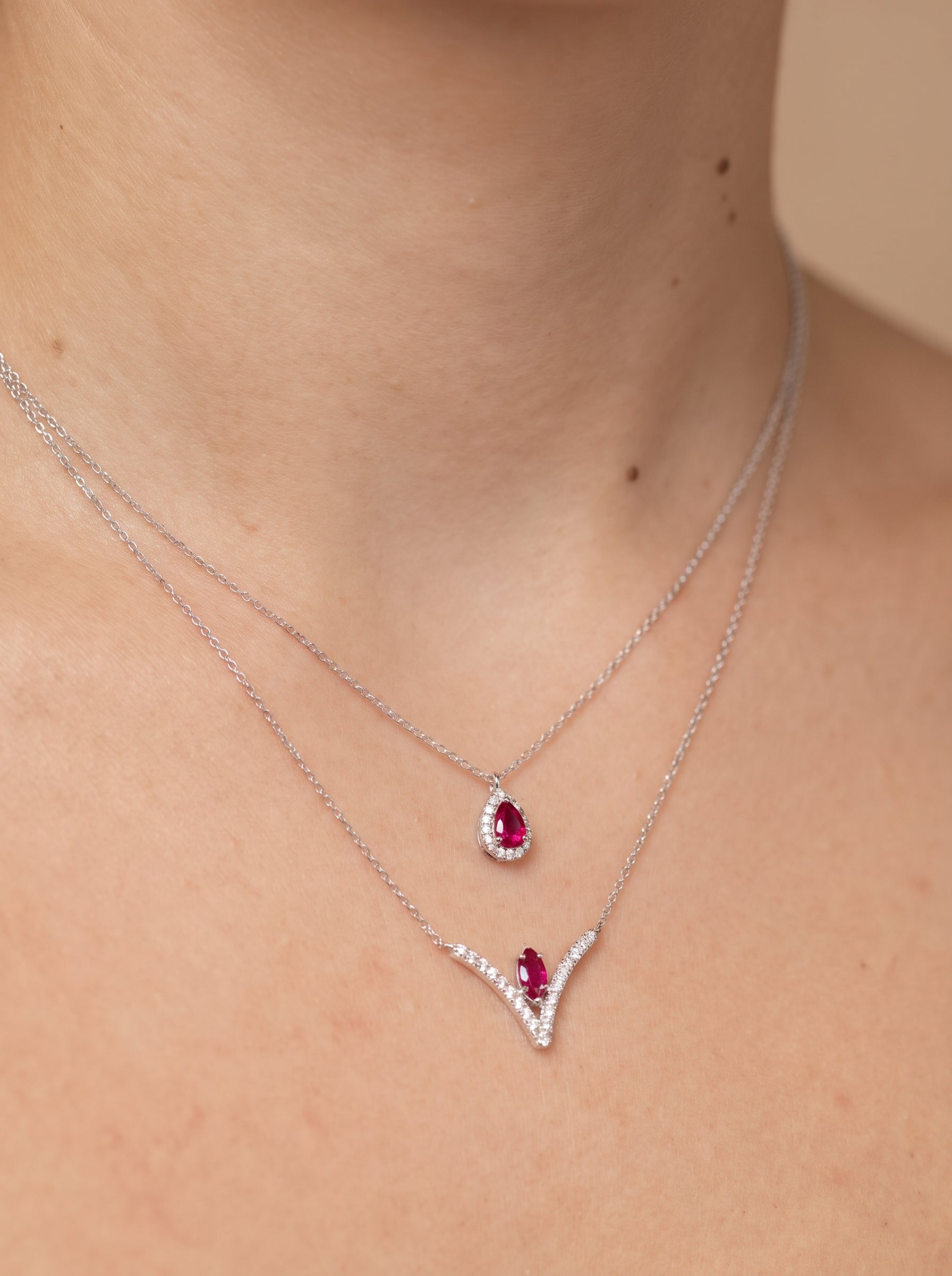 A close-up of a model's neck and collarbone wearing two layered silver necklaces. The shorter necklace features a teardrop-shaped ruby pendant, while the longer necklace has a V-shaped pendant with a small ruby and clear stones.