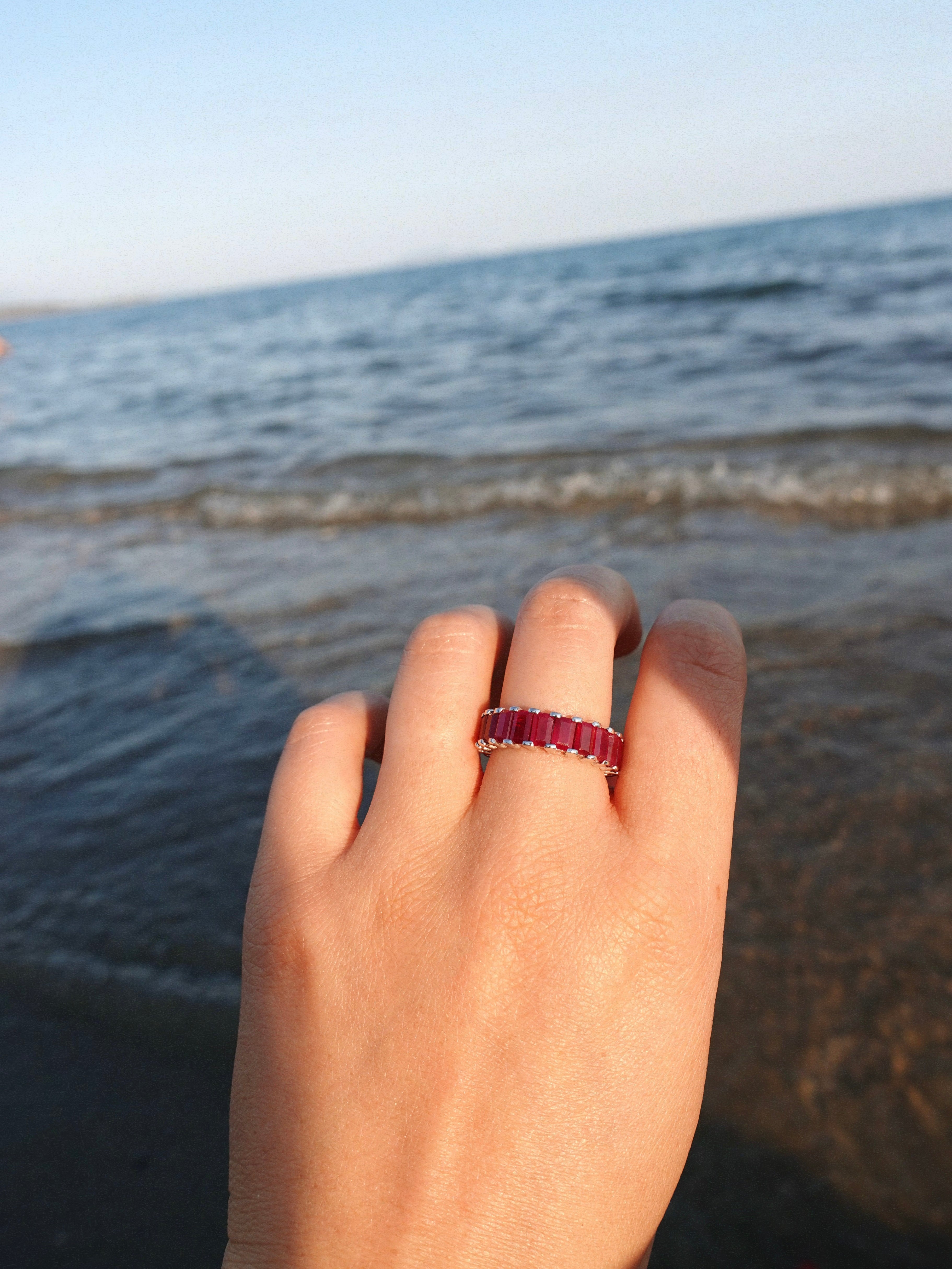 a hand wearing a ruby eternity ring over a background of sea waves