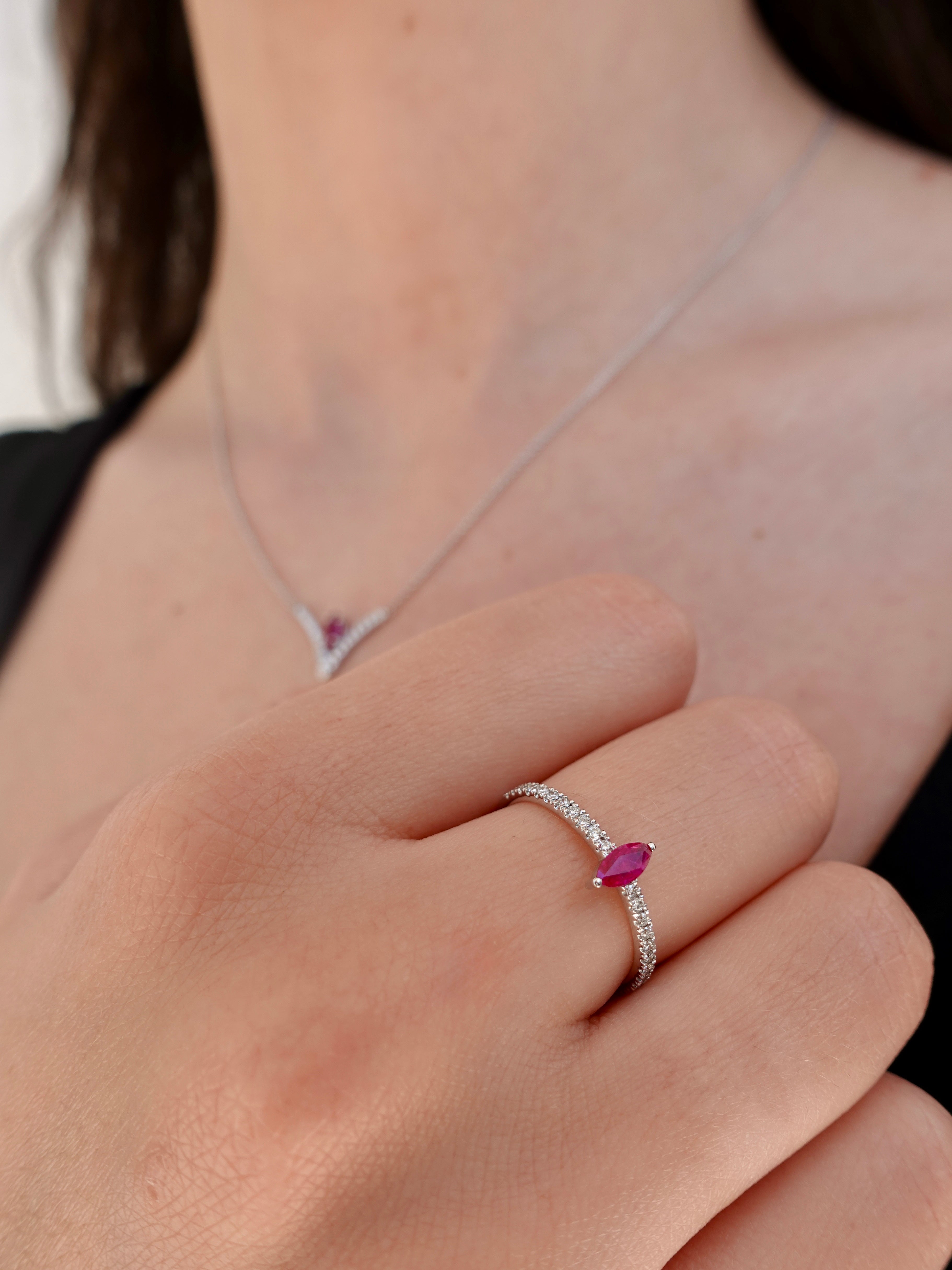 Close-up of a hand wearing a white-gold ring with a ruby gemstone, with a matching necklace in the background.