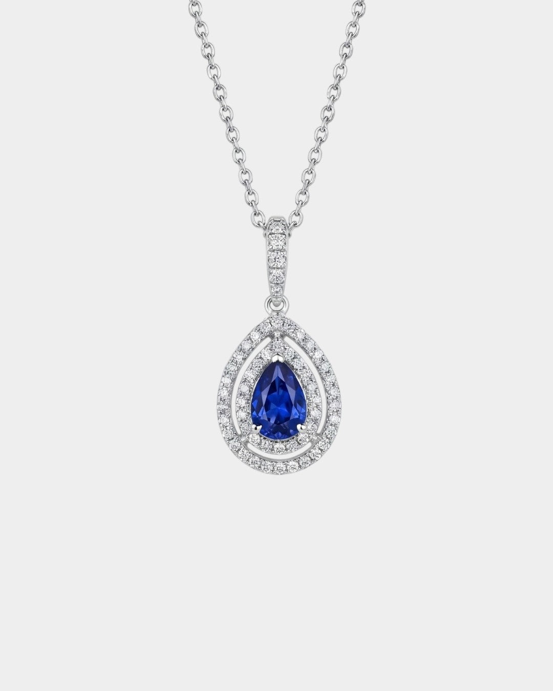 Gold necklace with a sapphire pendant and double diamond halo on a white background
