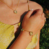 A close-up lifestyle shot of a woman wearing a matching gold jewelry set, including a delicate chain necklace with a small flower-shaped pendant and a bracelet with a matching charm, styled with a yellow floral top against a blurred green background.