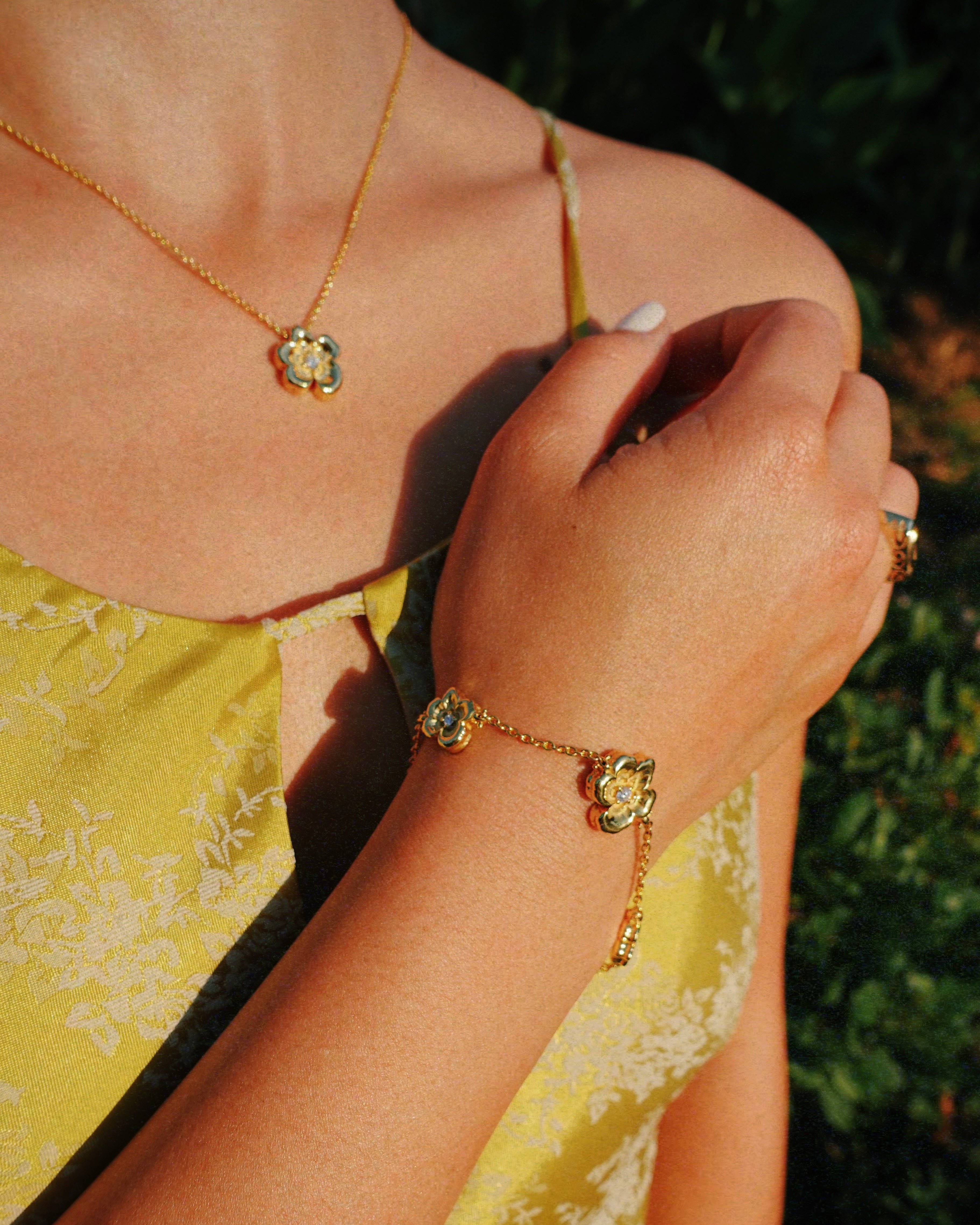 A close-up lifestyle shot of a woman wearing a matching gold jewelry set, including a delicate chain necklace with a small flower-shaped pendant and a bracelet with a matching charm, styled with a yellow floral top against a blurred green background.