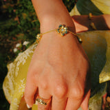 A gold chain bracelet with three flower charms, worn on a person's wrist, with a matching gold ring on their finger. The person is wearing a yellow dress, and the background shows a blurred outdoor garden setting.