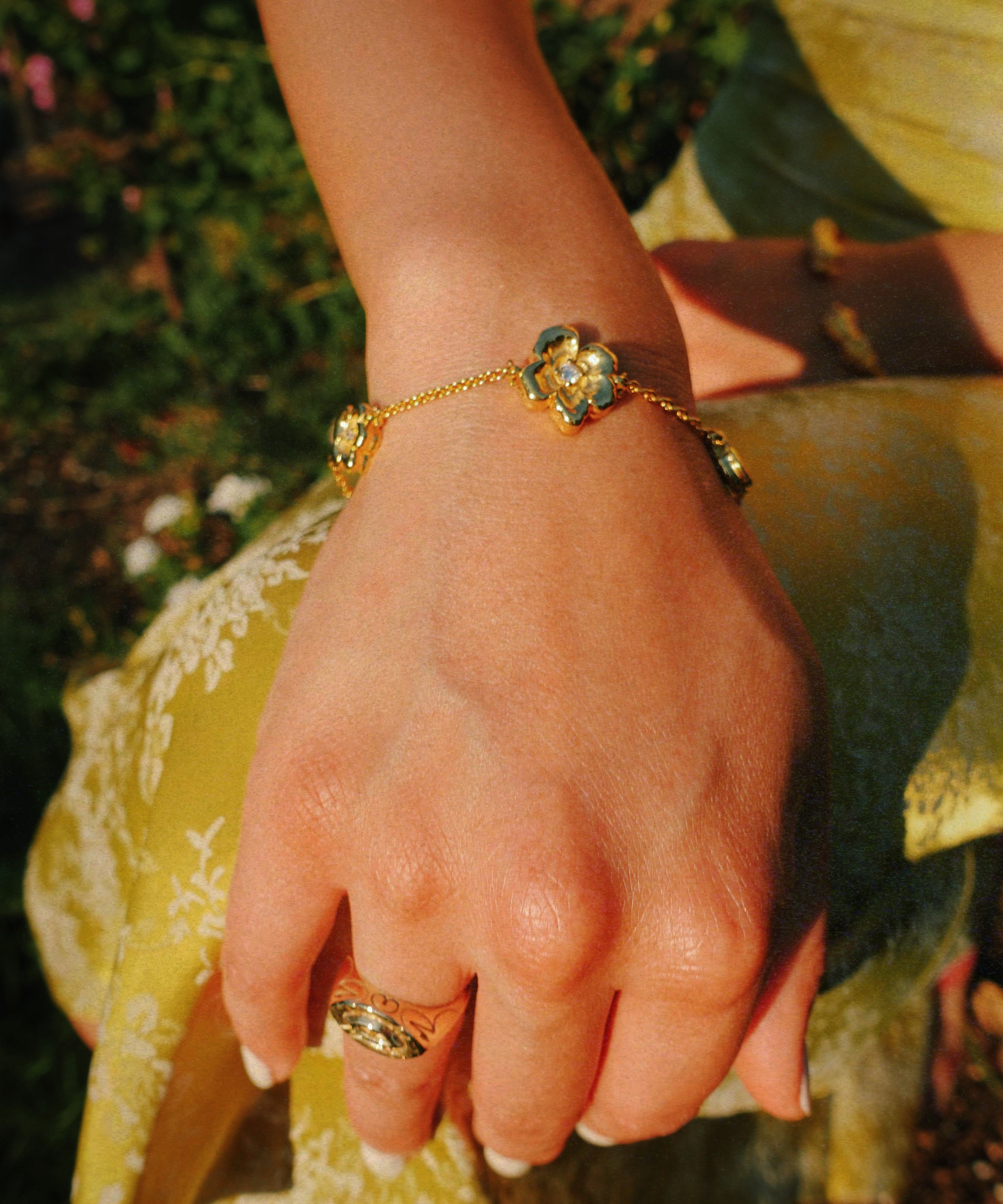 A gold chain bracelet with three flower charms, worn on a person's wrist, with a matching gold ring on their finger. The person is wearing a yellow dress, and the background shows a blurred outdoor garden setting.