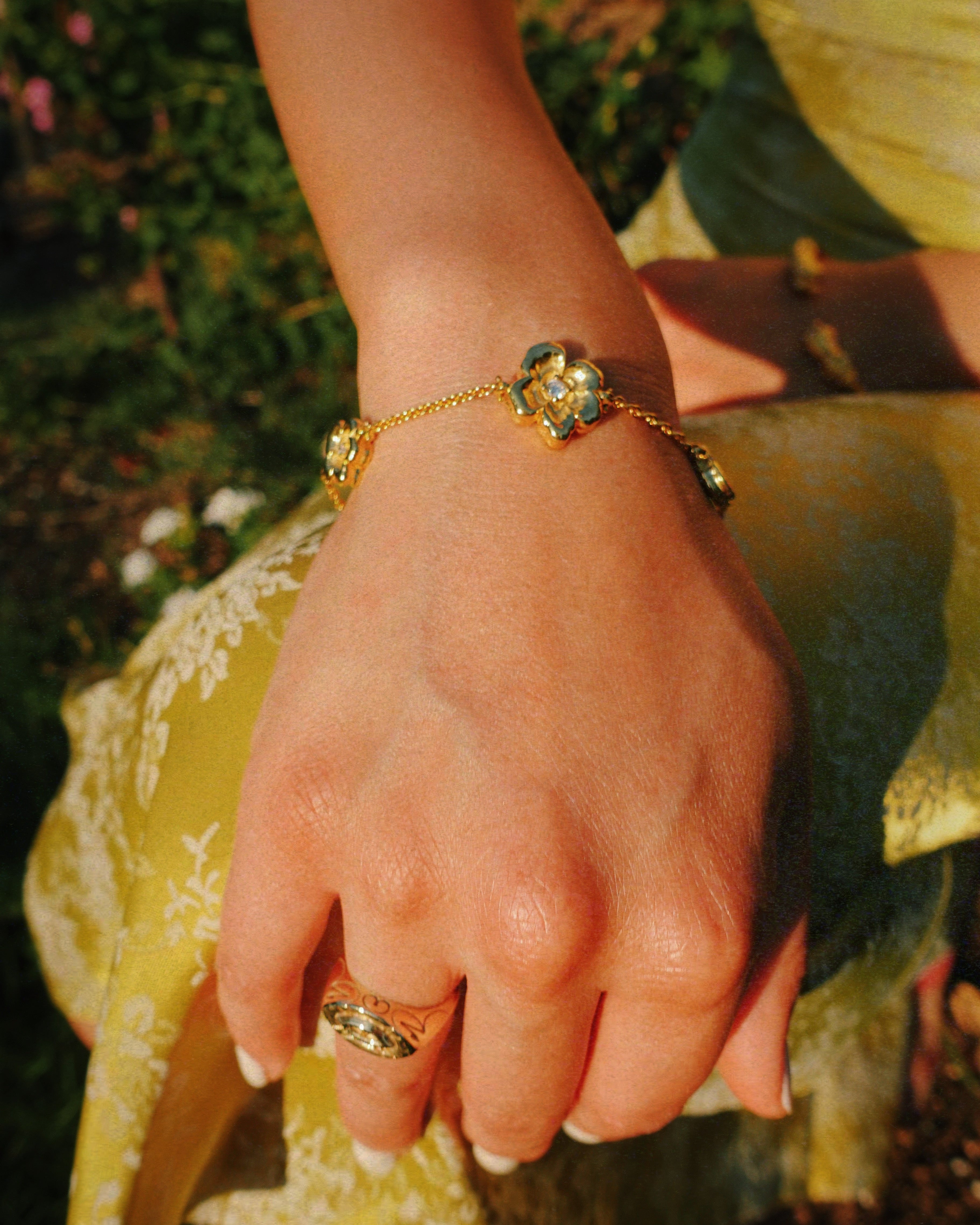 A gold chain bracelet with three flower charms, worn on a person's wrist, with a matching gold ring on their finger. The person is wearing a yellow dress, and the background shows a blurred outdoor garden setting.