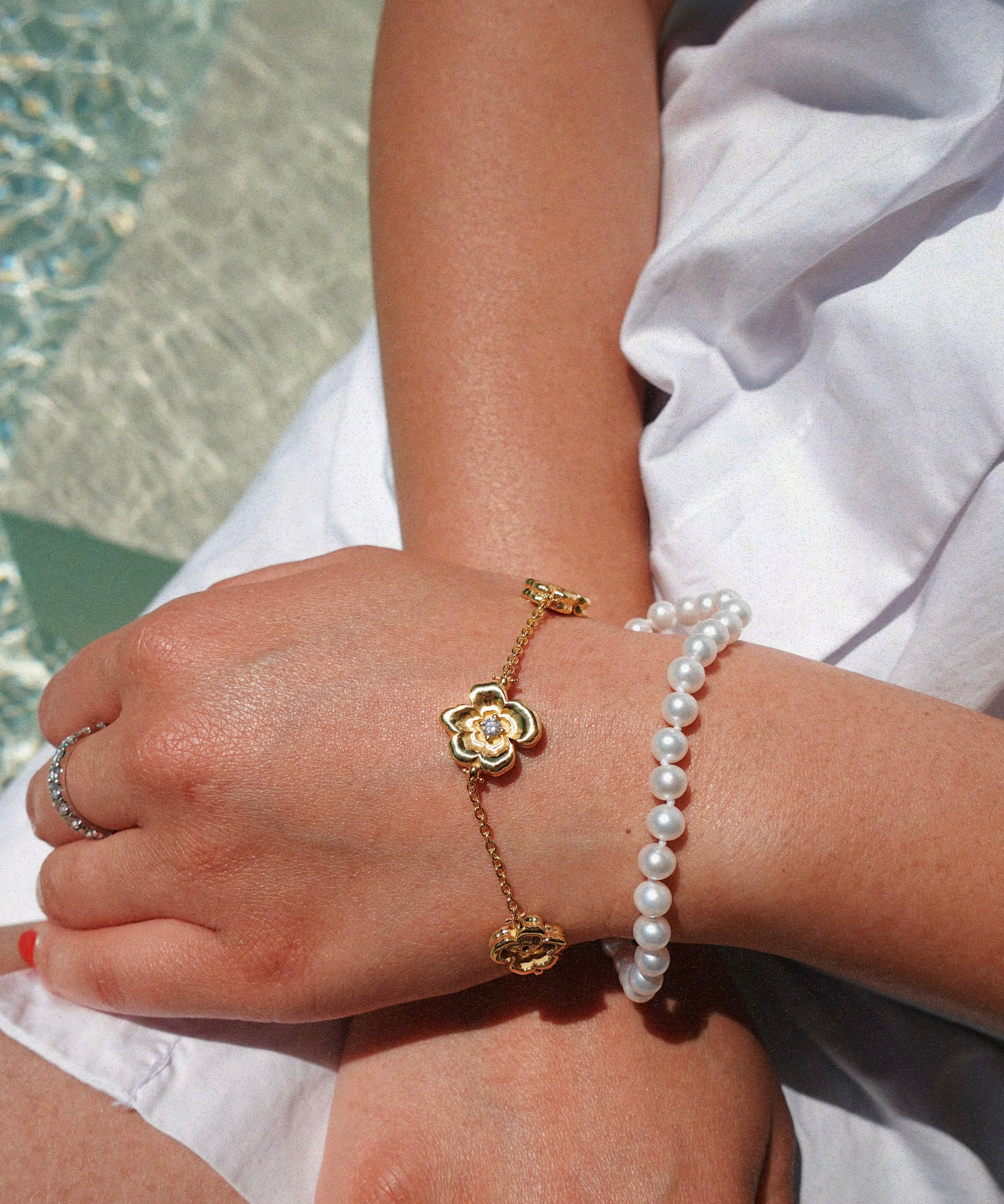 Close-up of a person's wrist wearing a gold flower bracelet and pearl bracelet by a pool.
