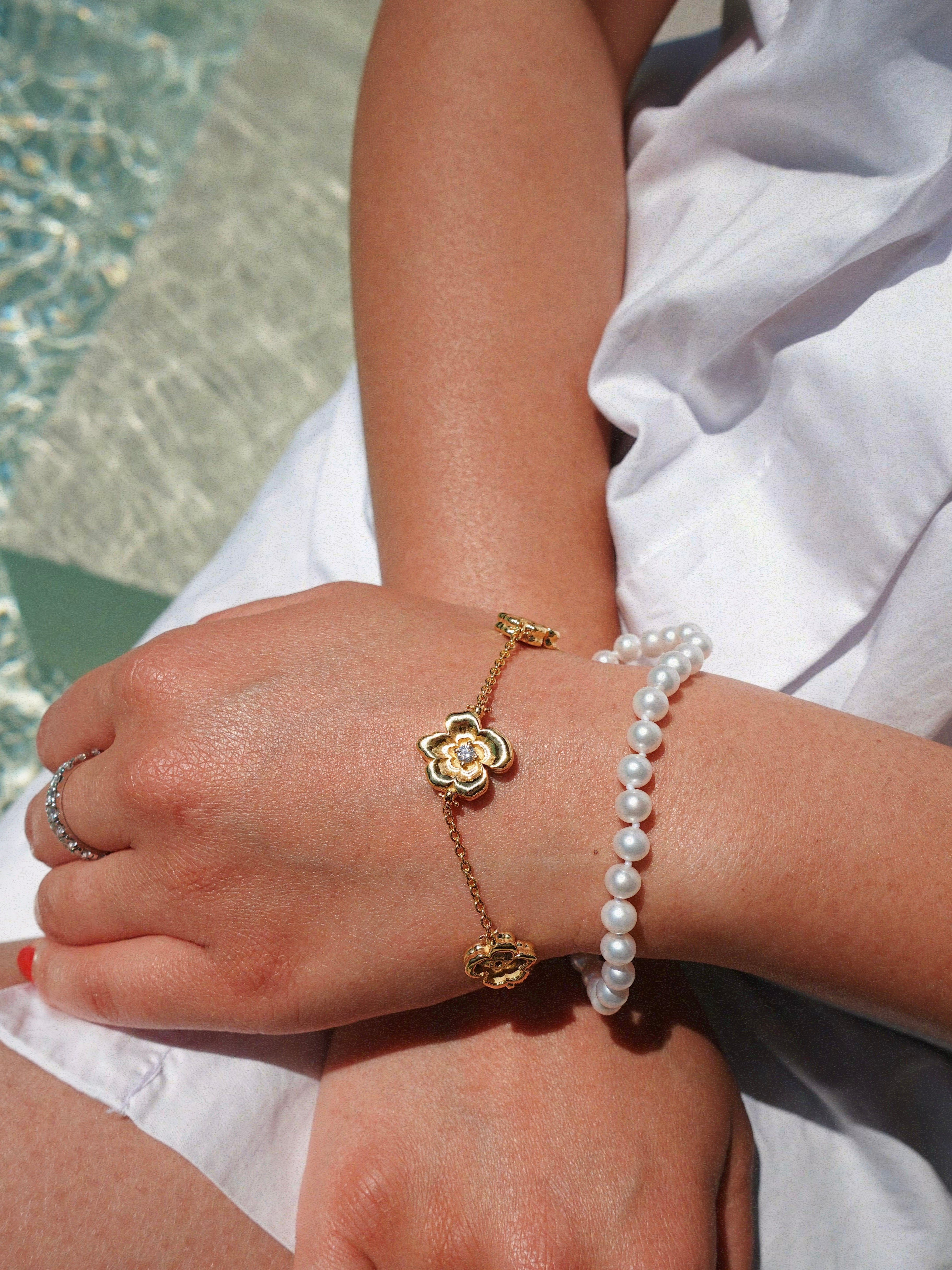 Close-up of a person's wrist wearing a gold flower bracelet and pearl bracelet by a pool.