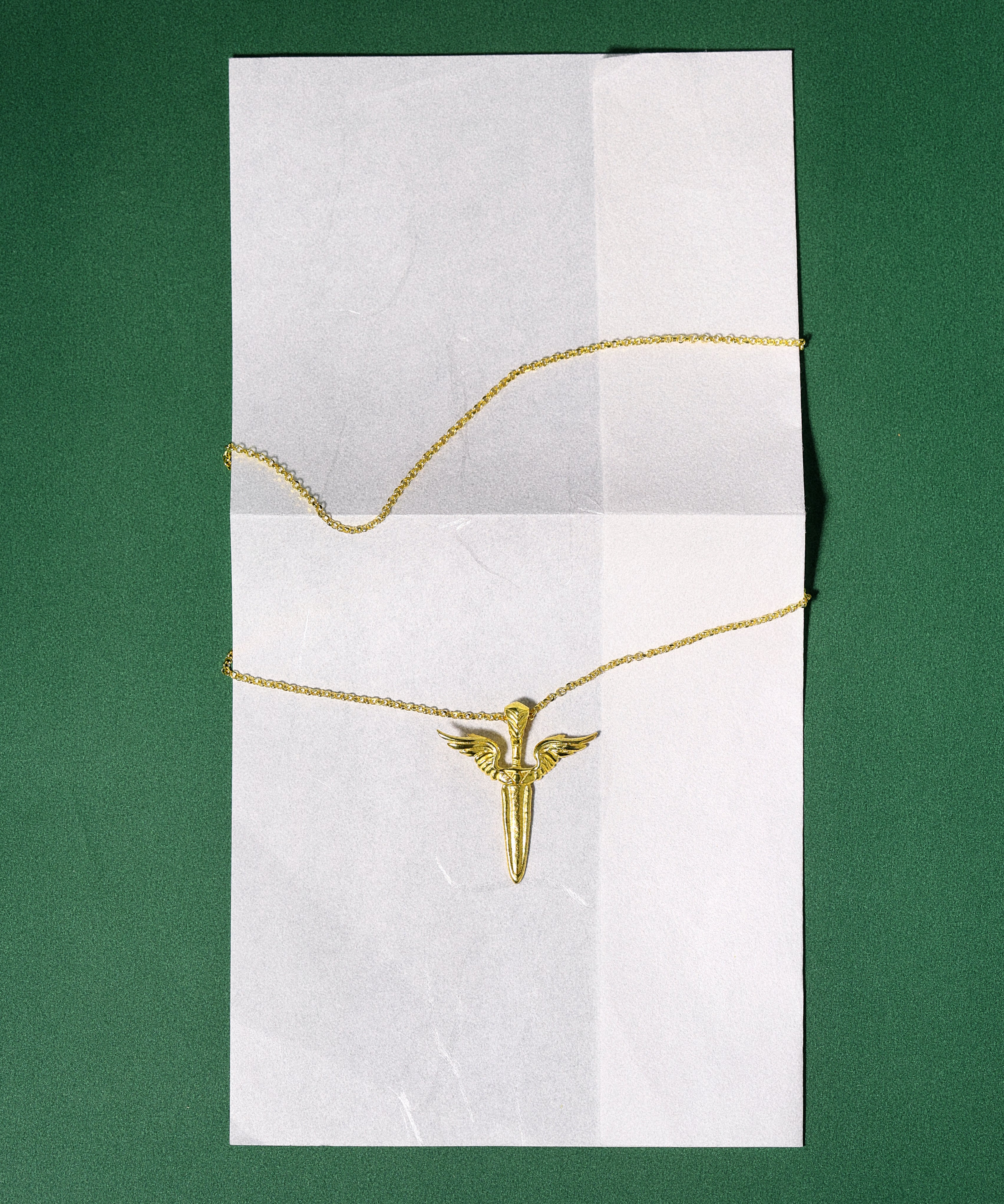 Gold necklace with a sword-shaped pendant on a white card against a green background