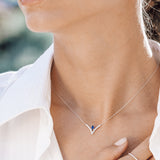 Close-up of a person wearing a silver necklace with a blue gemstone, set against a blurred outdoor background.