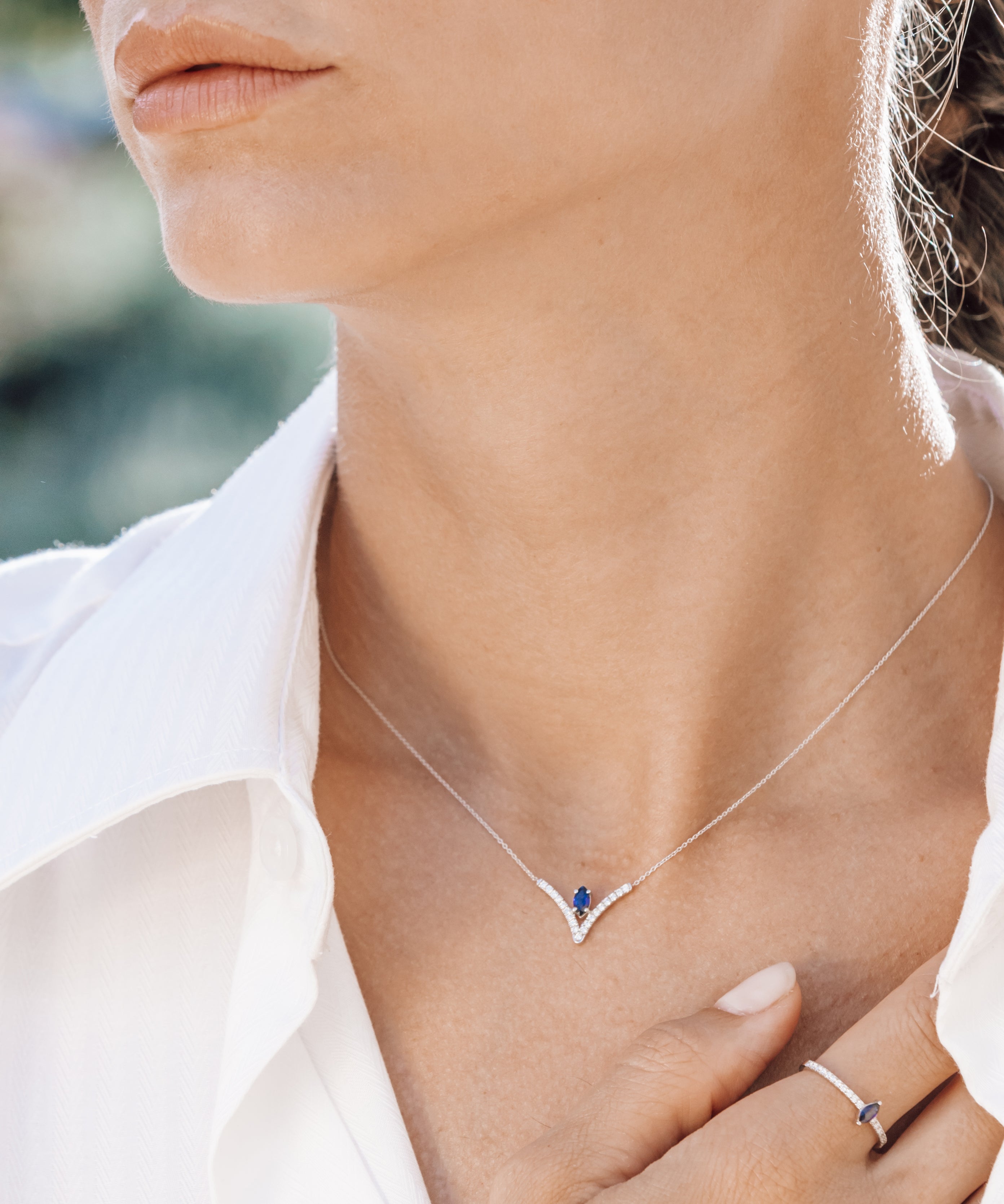 Close-up of a person wearing a silver necklace with a blue gemstone, set against a blurred outdoor background.