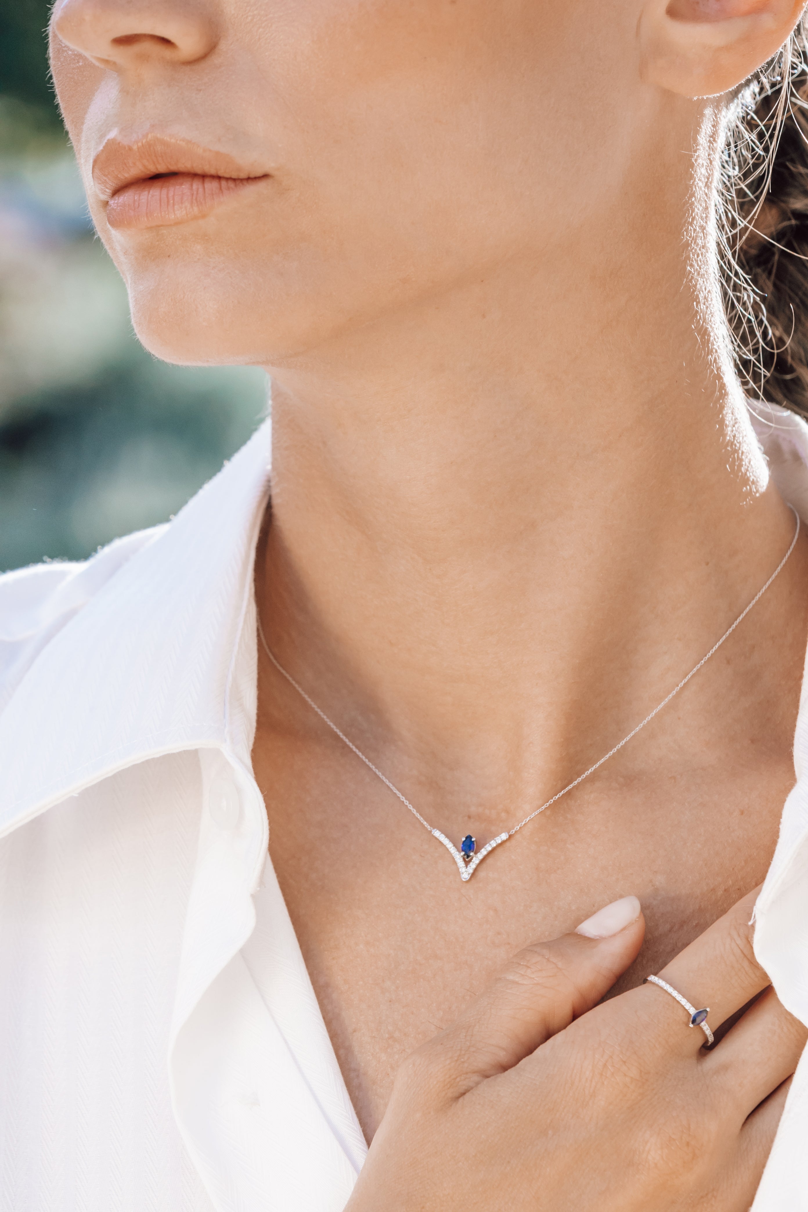 Close-up of a person wearing a silver necklace with a blue gemstone, set against a blurred outdoor background.